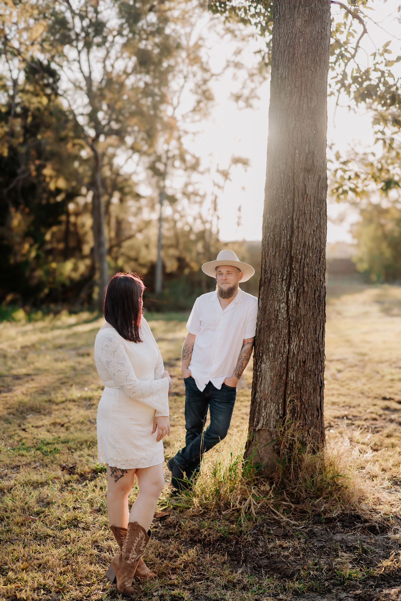 A couple shares a romantic moment near a tree in a sunlit field, woman in white dress and man in cowboy hat.