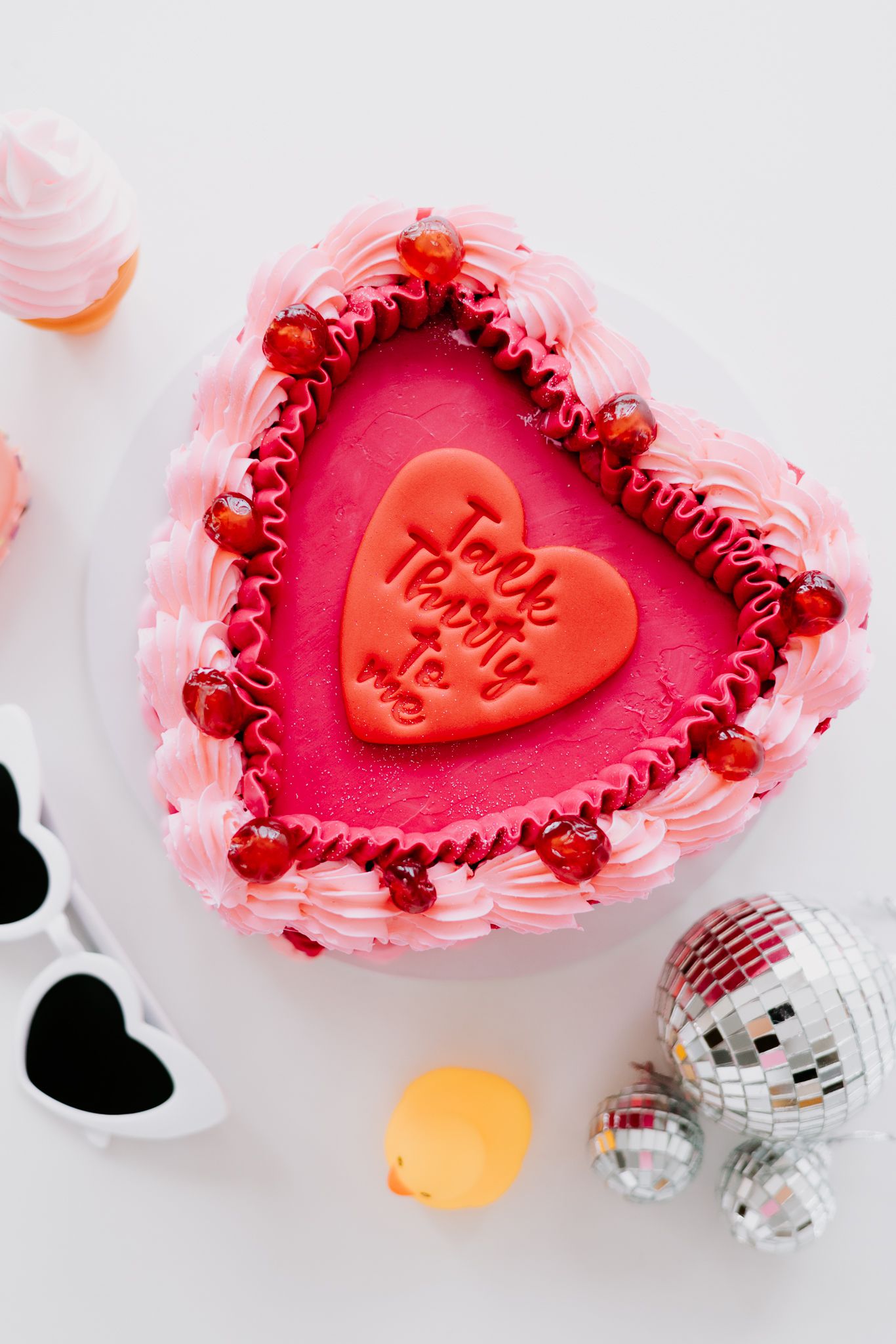 Heart-shaped pink cake with red fondant center, cherry decorations, disco balls, and heart sunglasses on white background.