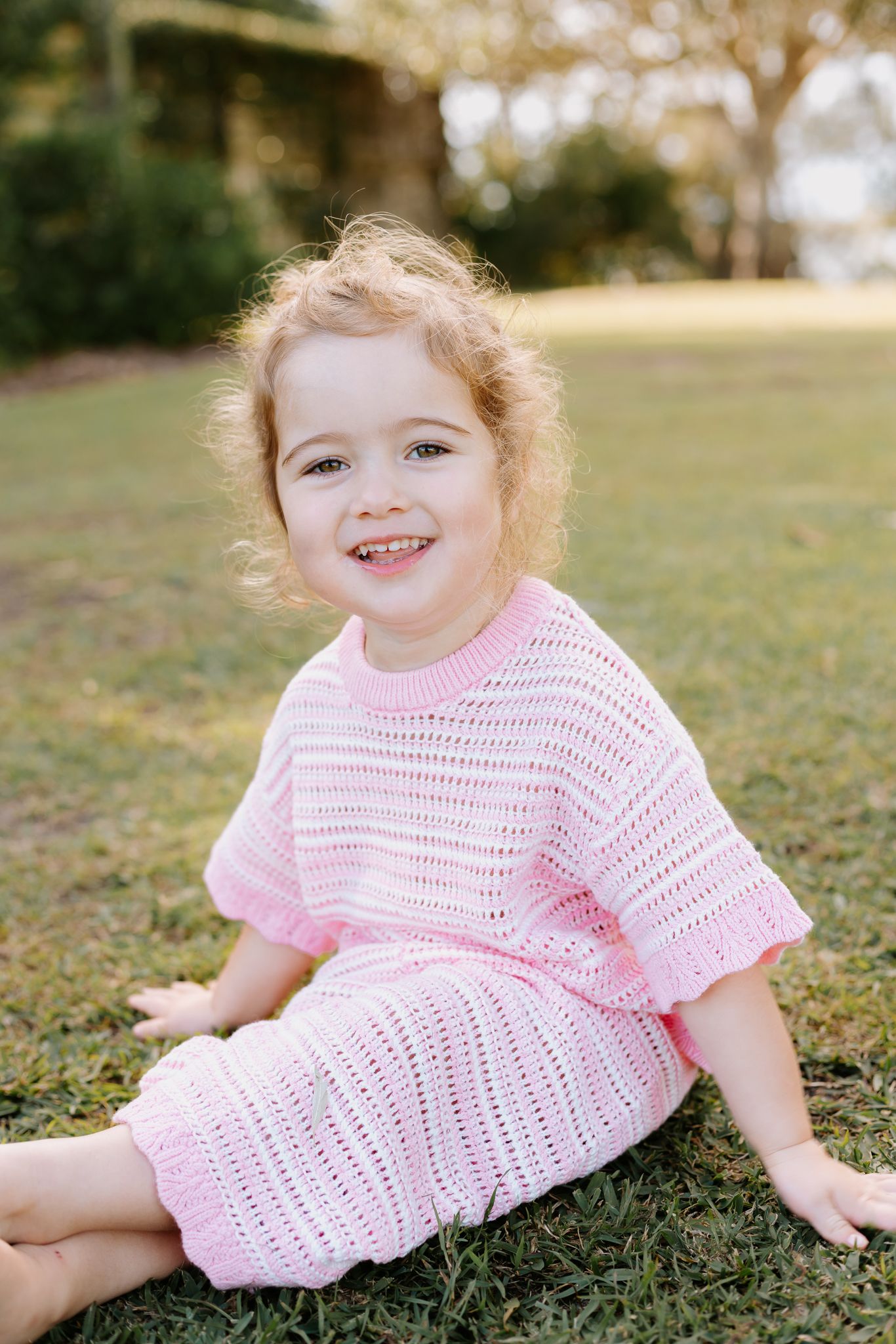 Toddler girl in pink knit outfit sitting on green grass, smiling cheerfully outdoors.