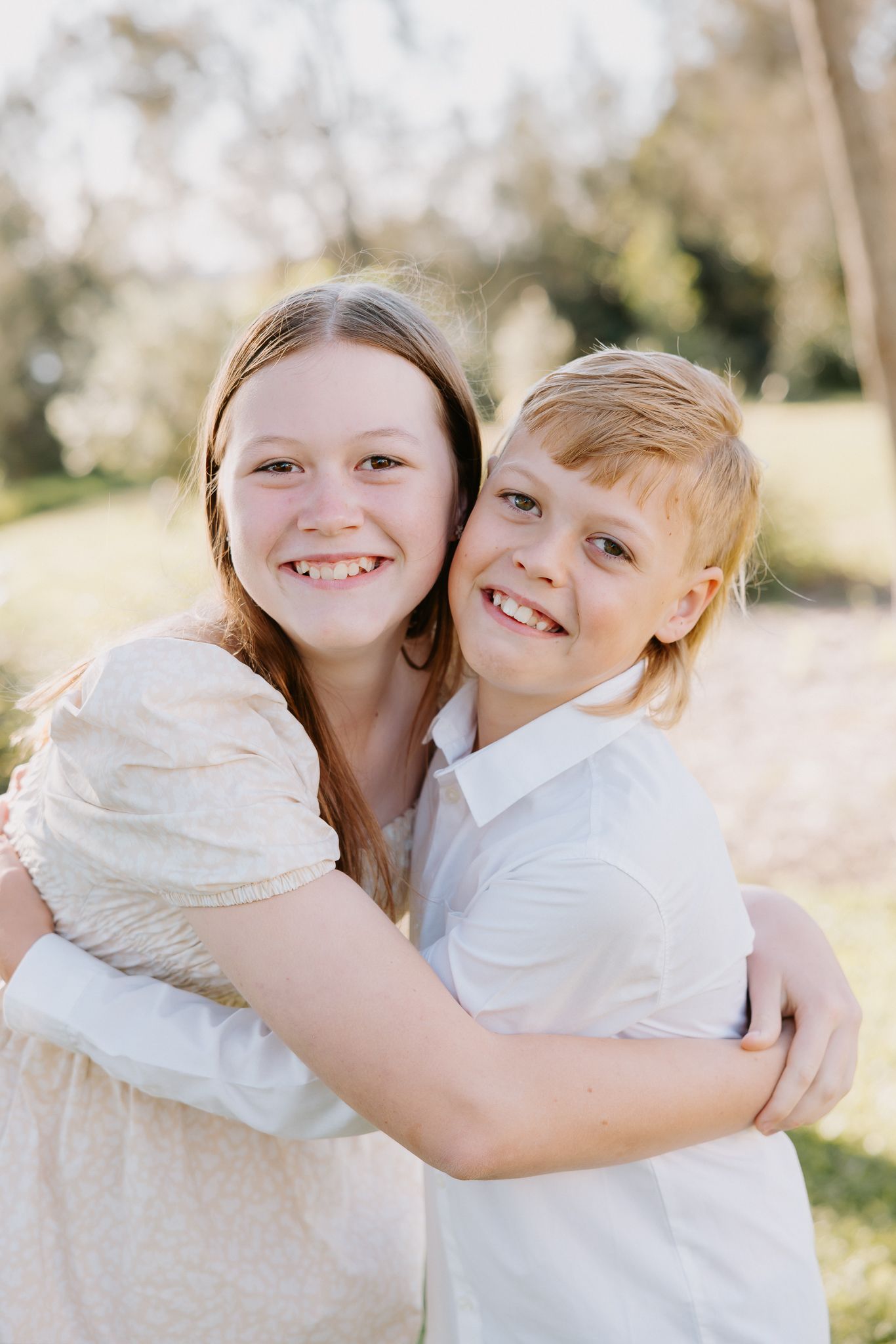 Two smiling children hugging outdoors in a sunny park, wearing white and cream outfits.