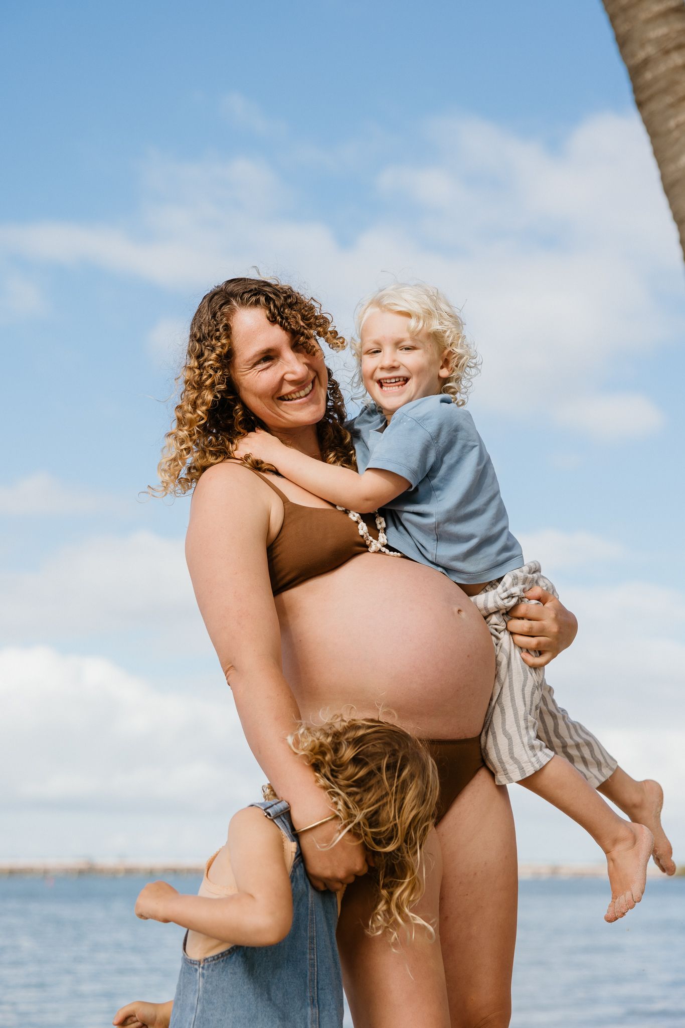 Smiling pregnant woman in brown bikini holding toddler at beach, with another child hugging her baby bump on sunny day.