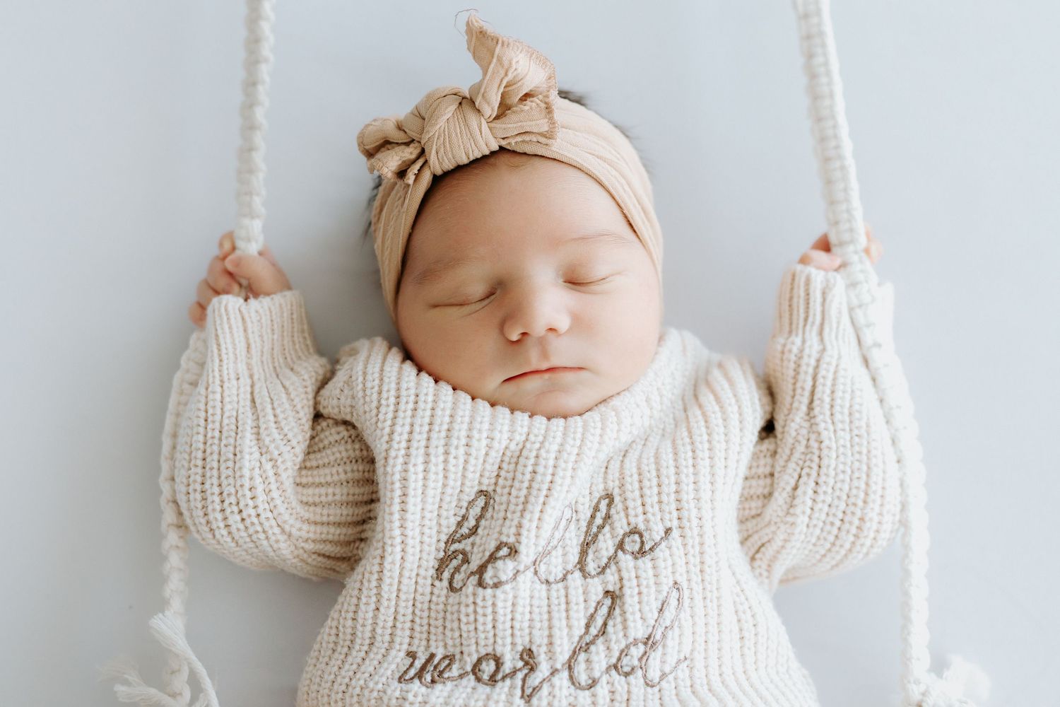 Sleeping newborn baby in cream knit sweater reading hello world with beige bow headband on a white swing.