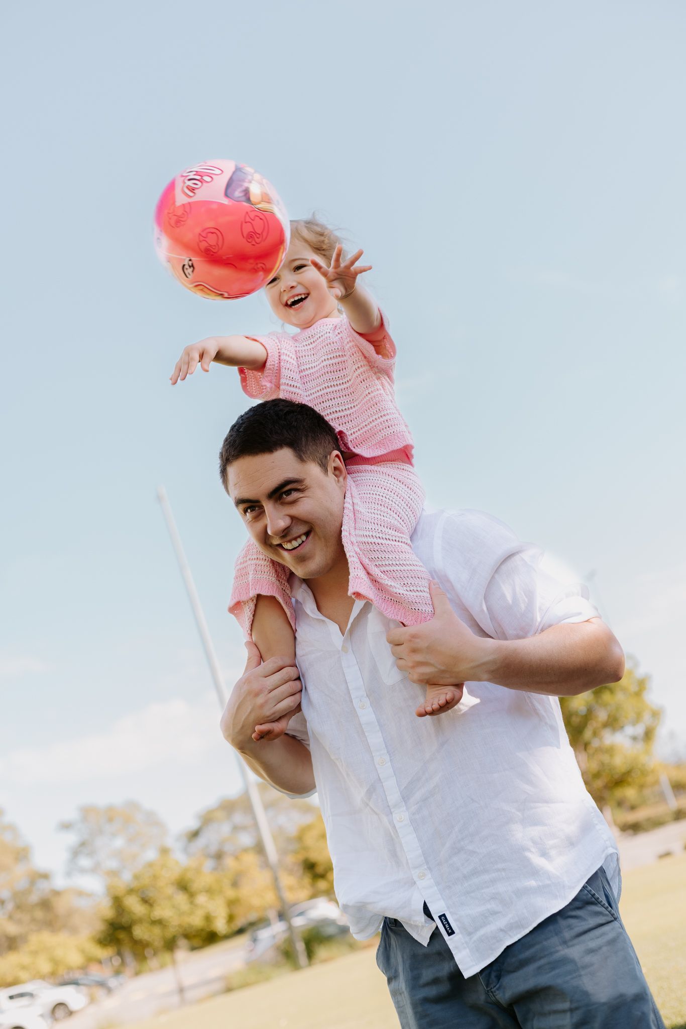 A smiling man carries a laughing toddler girl on his shoulders outdoors, as she reaches for pink balloons.
