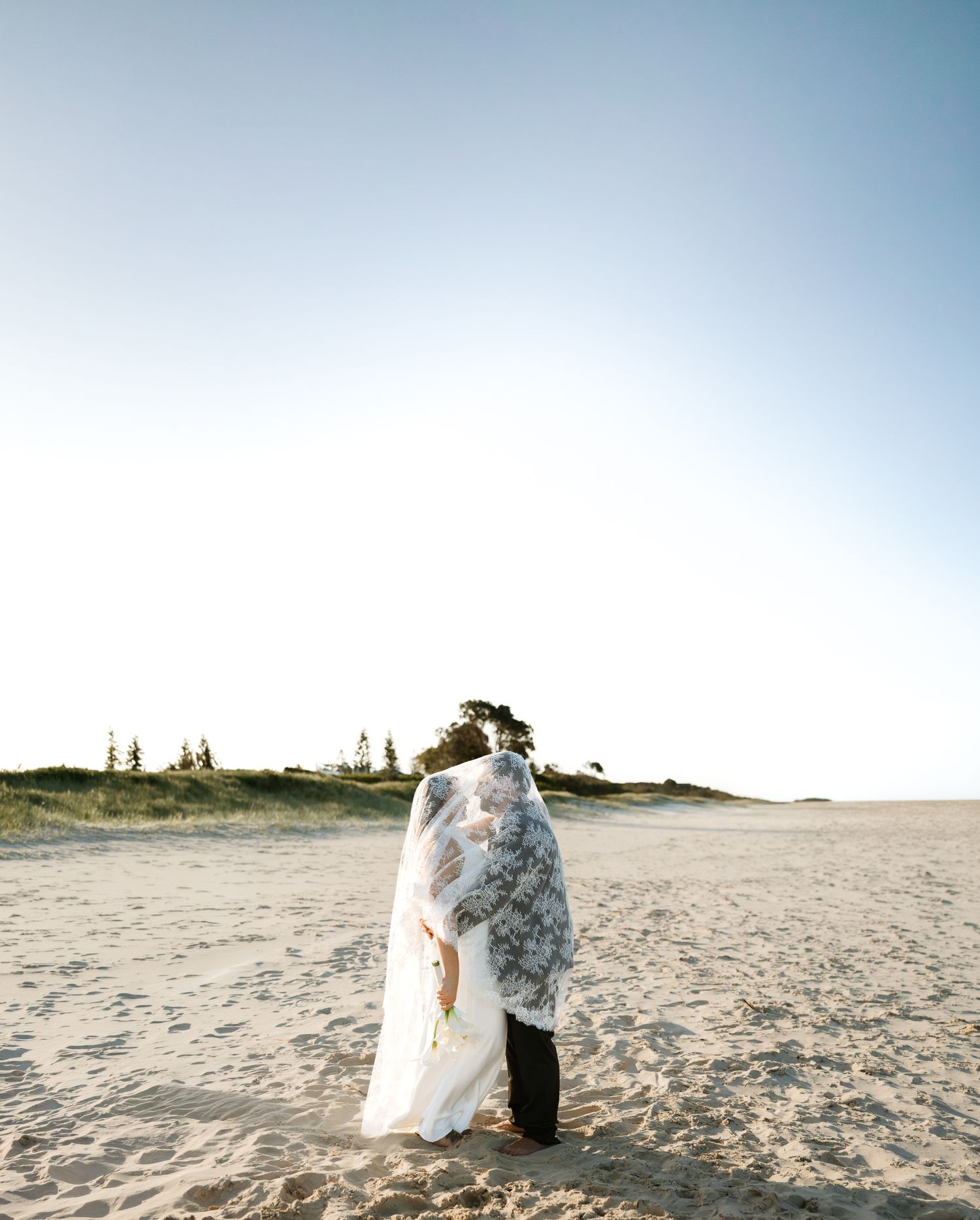 A couple shares a romantic kiss on a sandy beach, draped in a floral veil, with dunes and trees in the background.