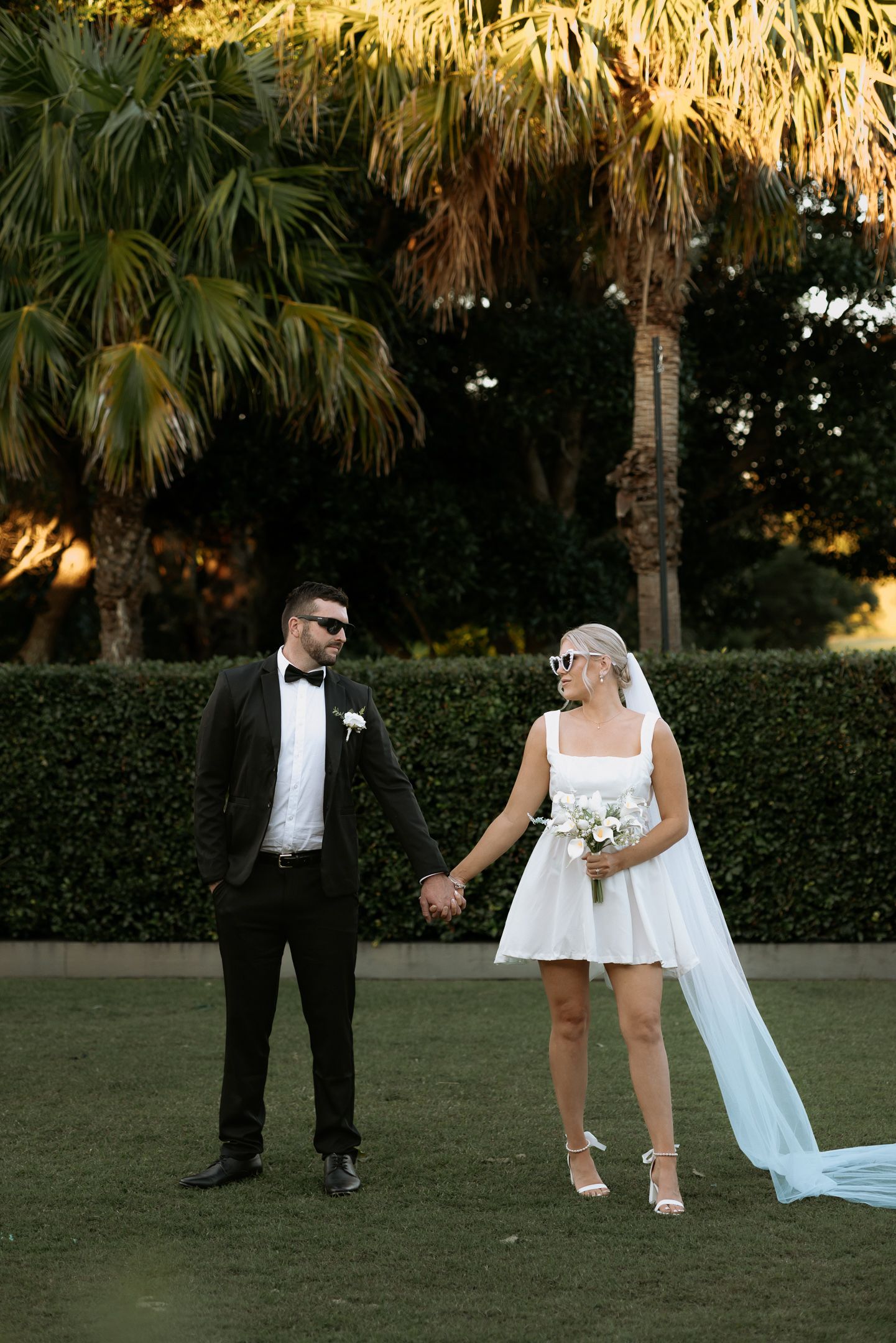 Bride in short white dress and groom in black tuxedo holding hands on green lawn with palm trees at sunset.