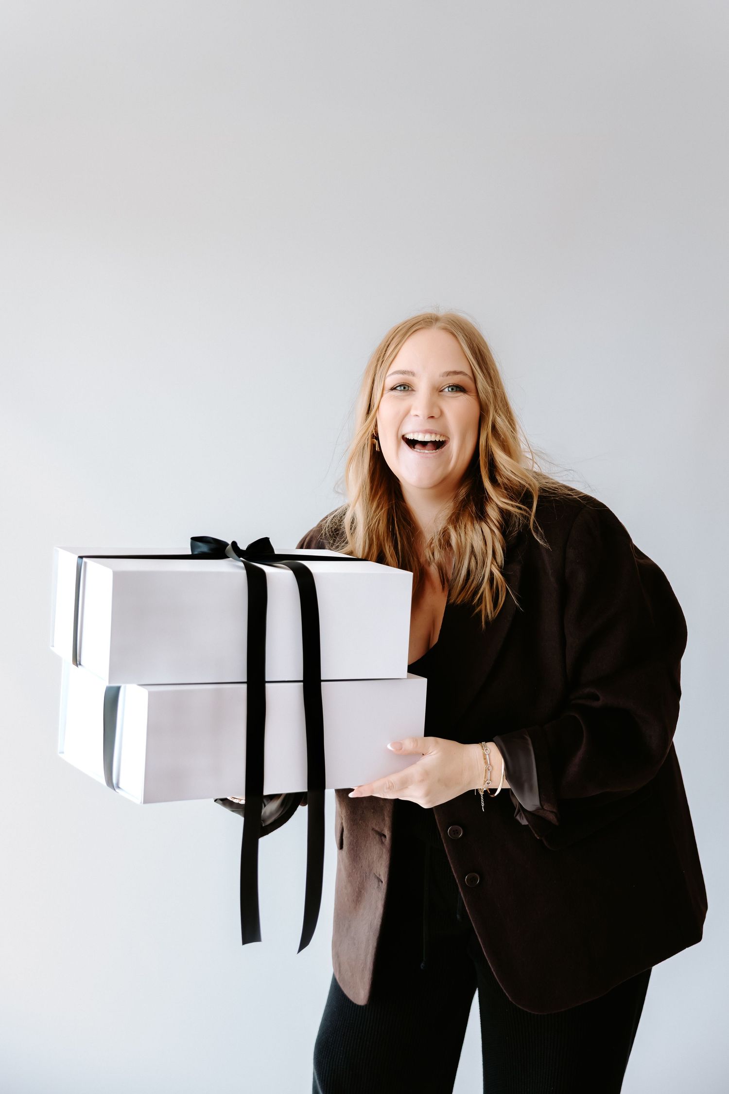 Smiling blonde woman in black blazer holding stacked white gift boxes with black ribbon bows against gray background.