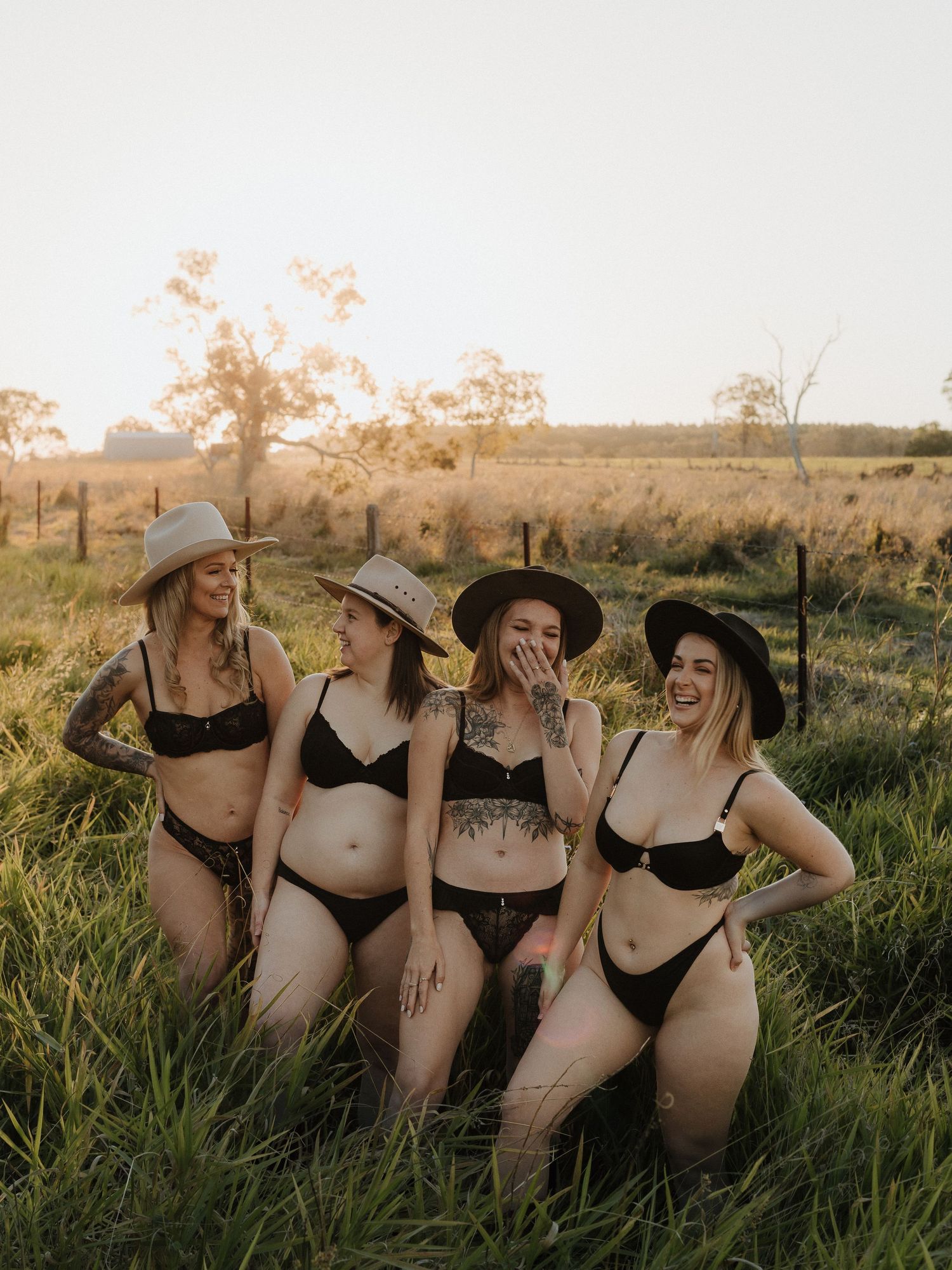 Four women in matching black lingerie and wide-brim hats laughing together in a sunny rural field.