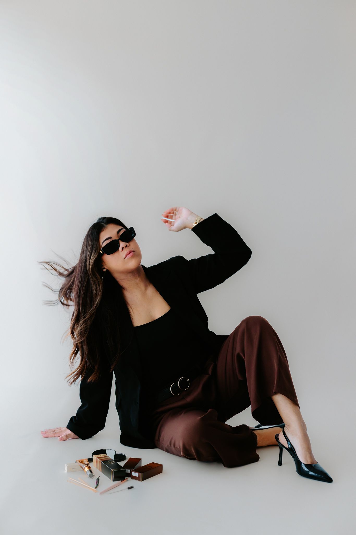 Stylish woman in black blazer and brown pants sitting on floor with sunglasses and heels in a studio setting.