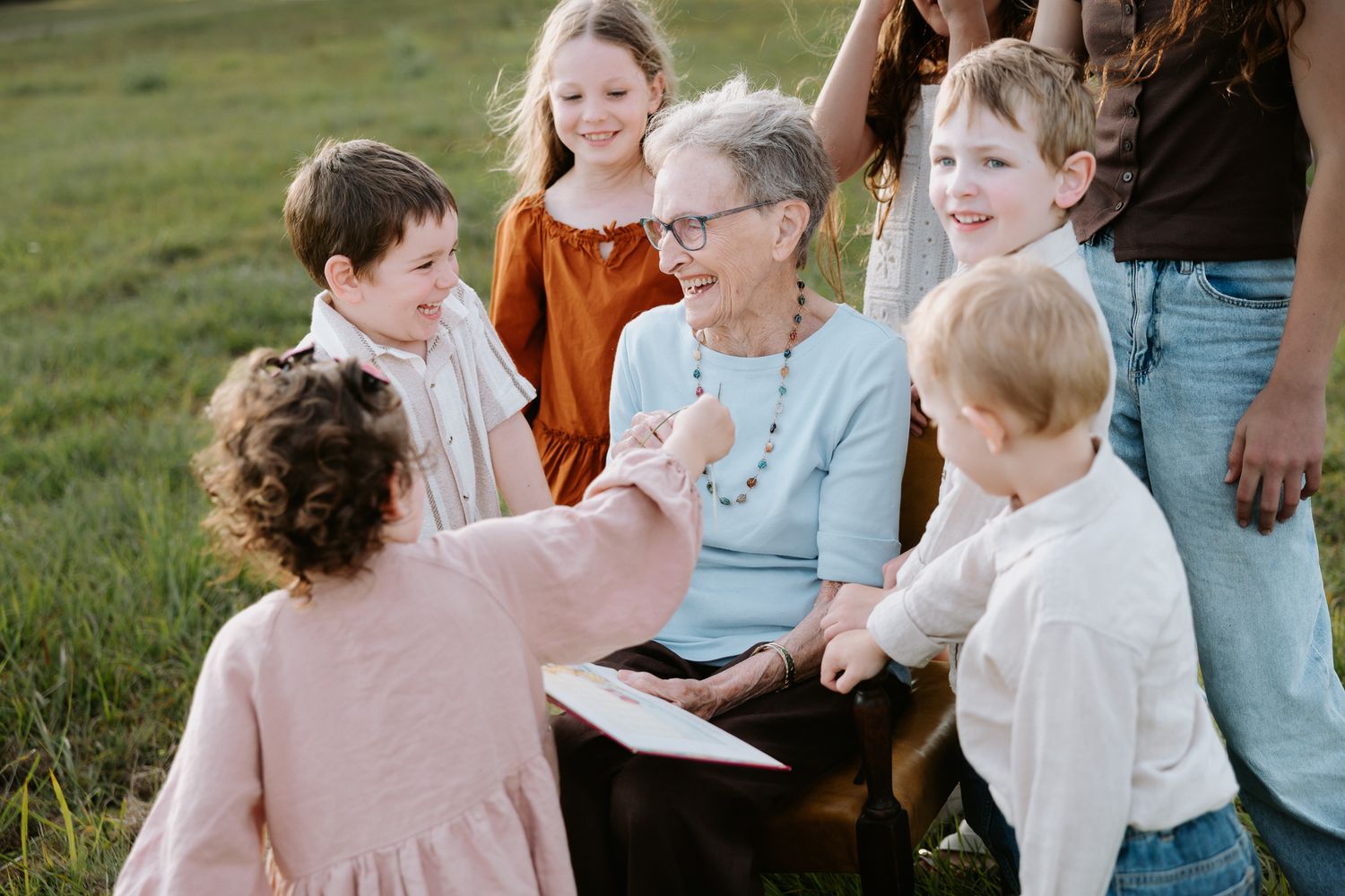 Elderly grandmother smiles warmly surrounded by young grandchildren outdoors in a green field during a family gathering.