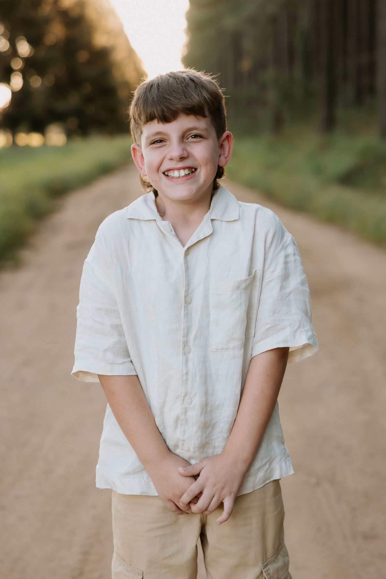 Smiling boy in white linen shirt and khaki shorts standing on a dirt path surrounded by trees in golden light.