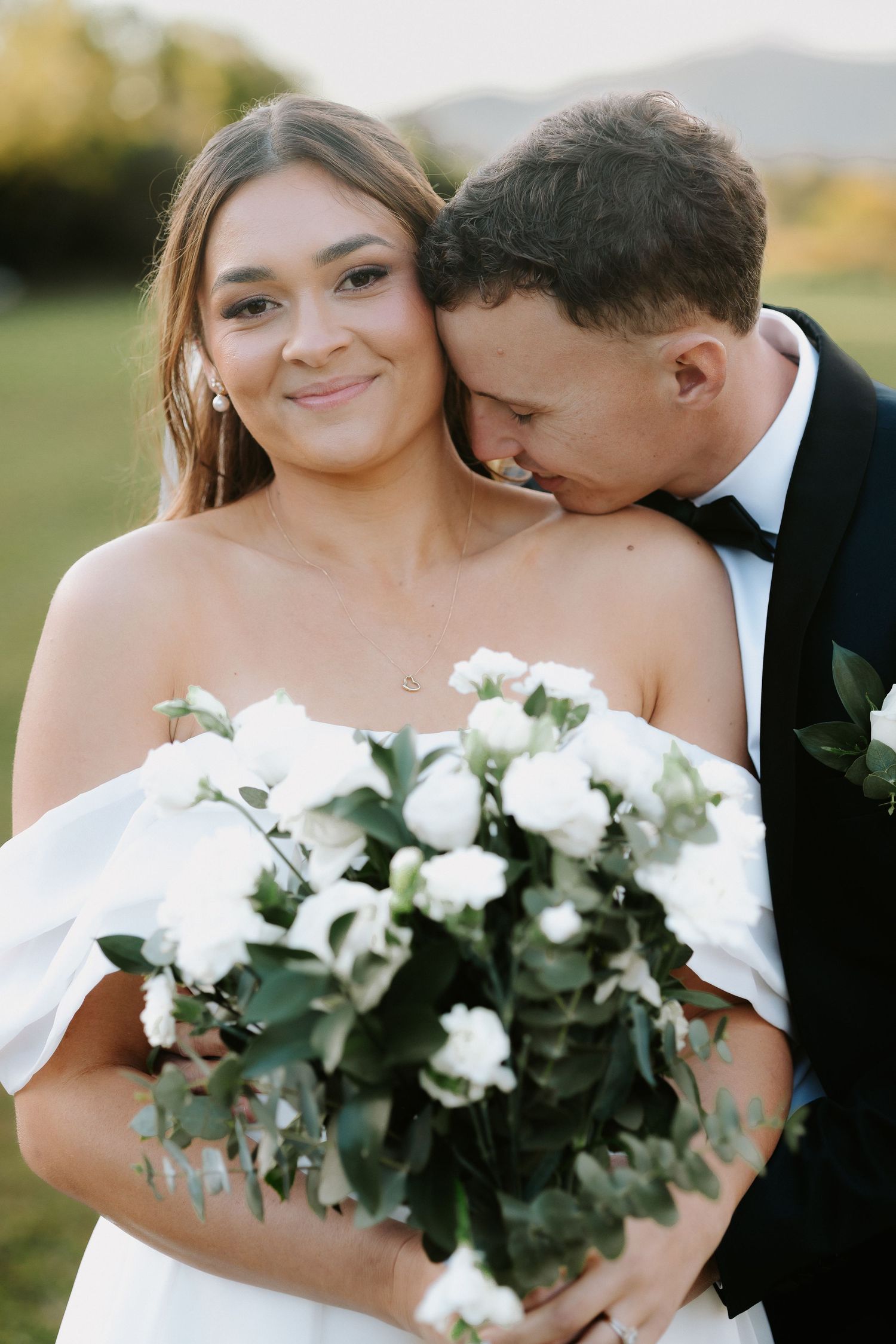Bride holding white rose bouquet smiles as groom in black tuxedo kisses her neck outdoors at golden hour.