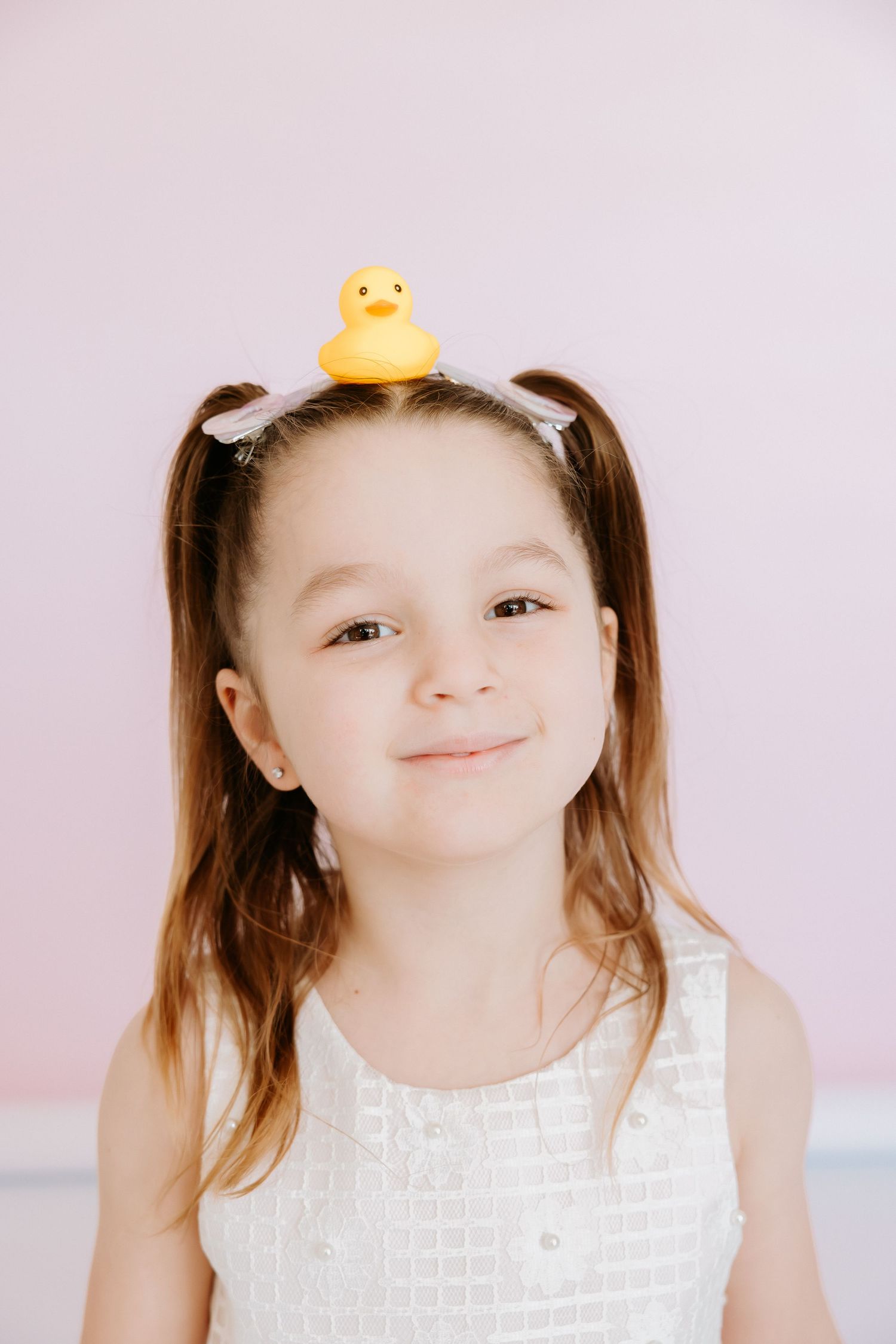 Young girl with pigtails balancing a yellow rubber duck on her head, wearing a white dress against a pink background.