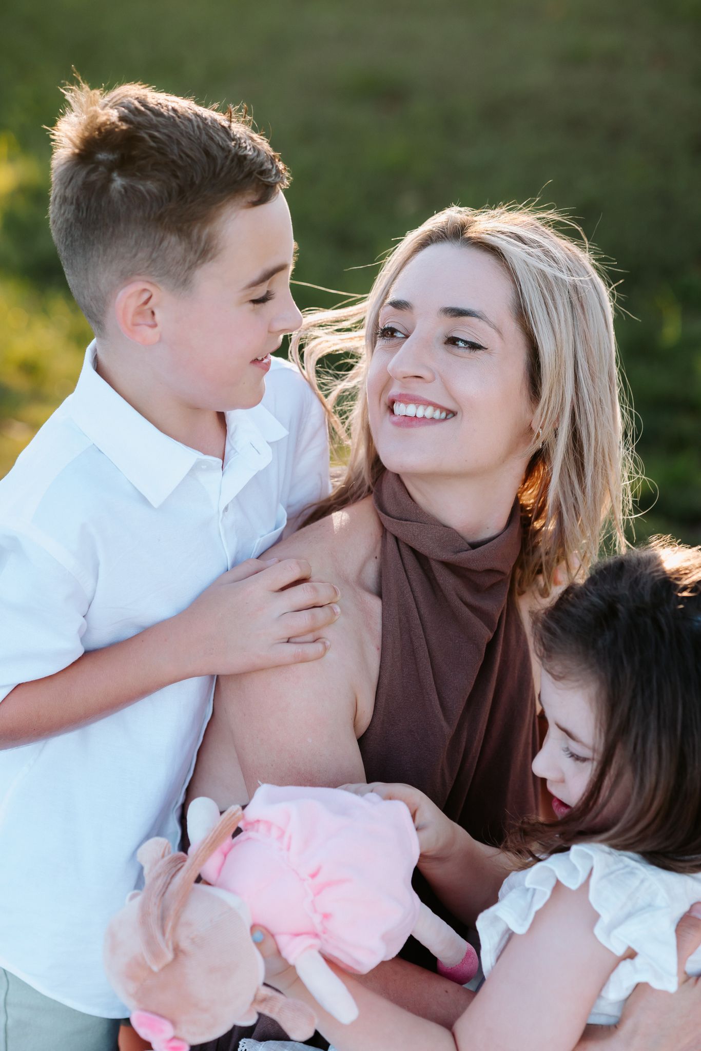 A smiling blonde mother hugs her son and daughter outdoors in a sunny green field.