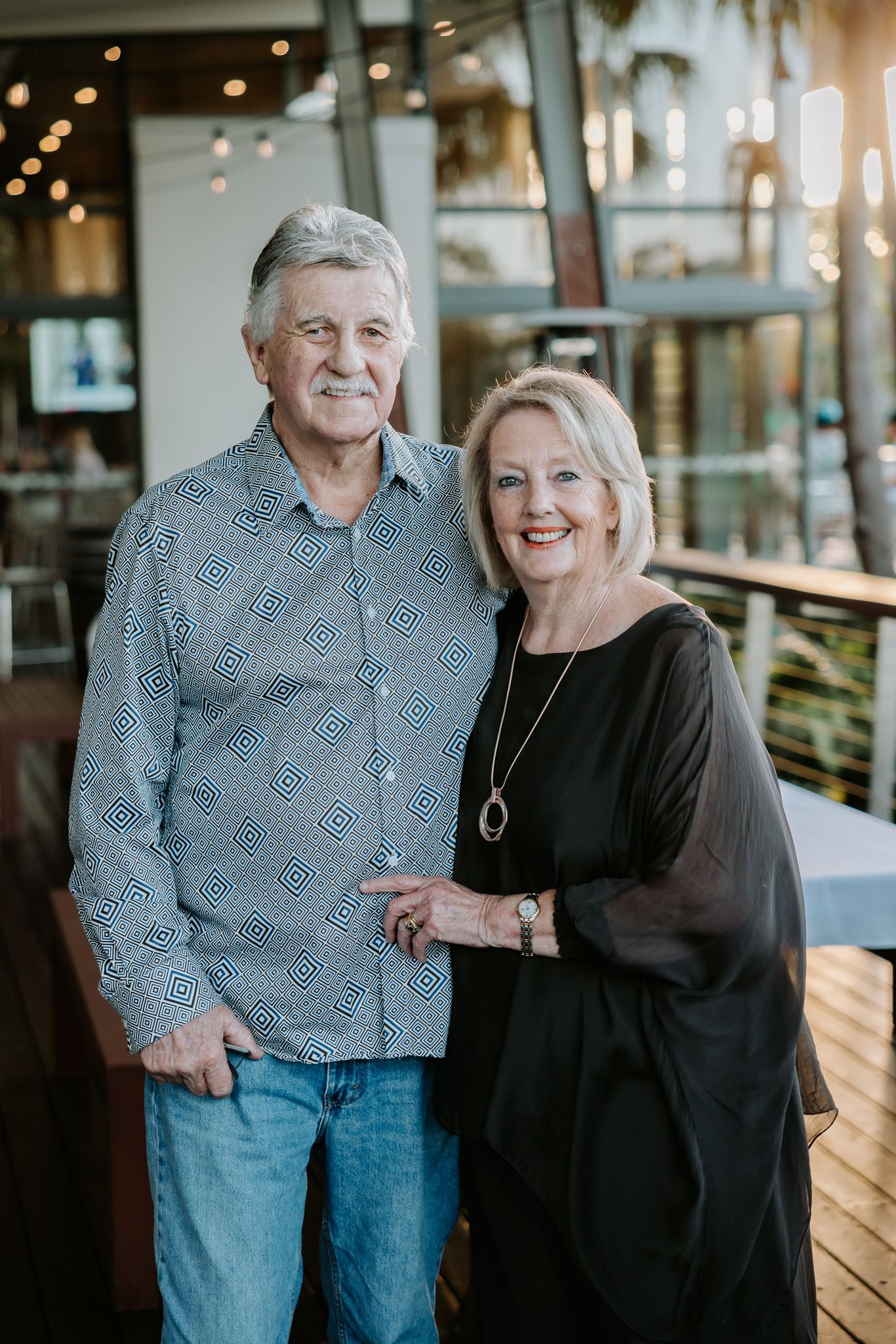 Smiling older couple posing together on a restaurant patio, man in patterned shirt and jeans, woman in black dress.