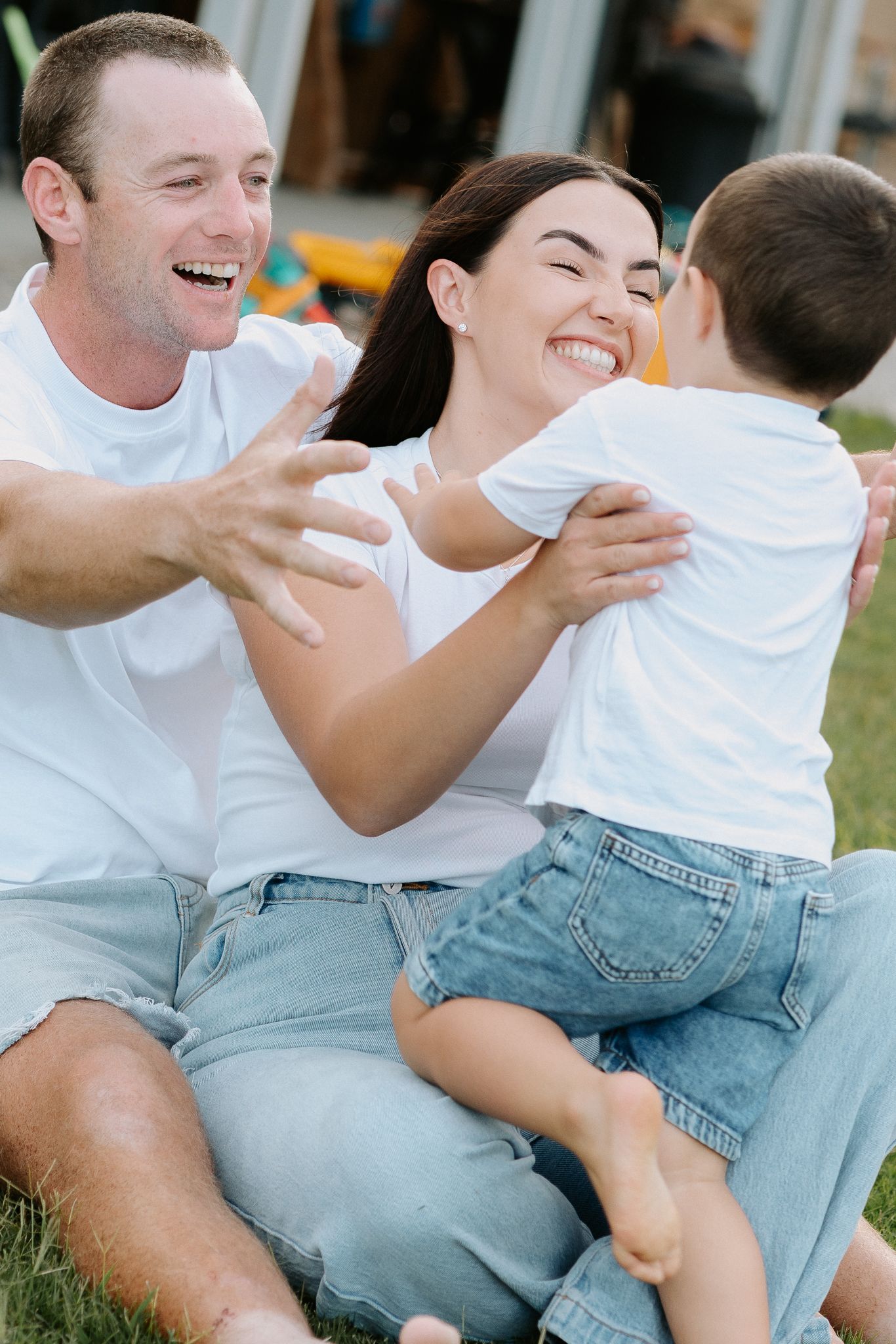 Happy family in white tees and jeans sitting on grass, parents laughing as child climbs into mother's arms.