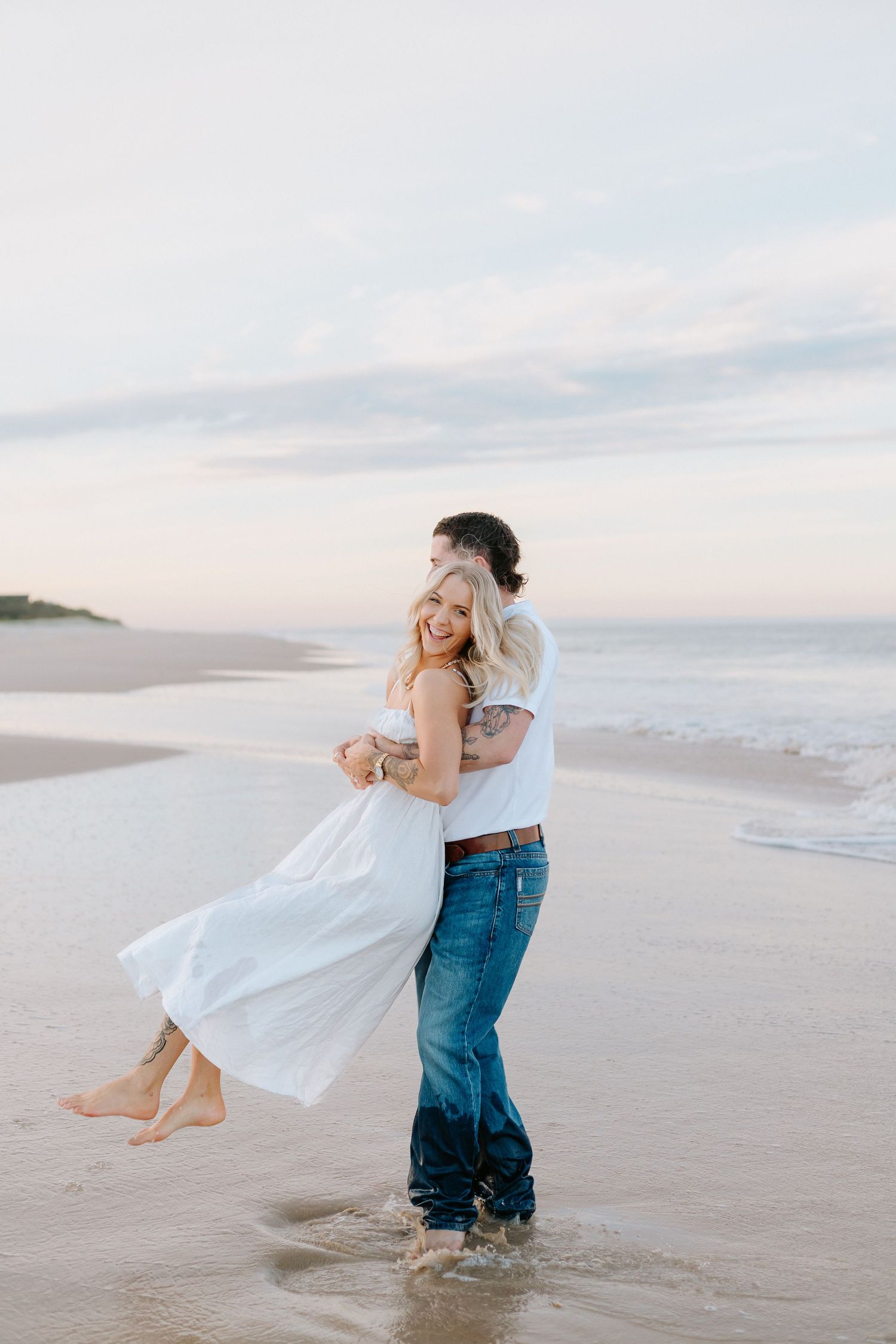 Couple embracing on a sandy beach at sunset, woman in white dress lifted by man in jeans and white tee.