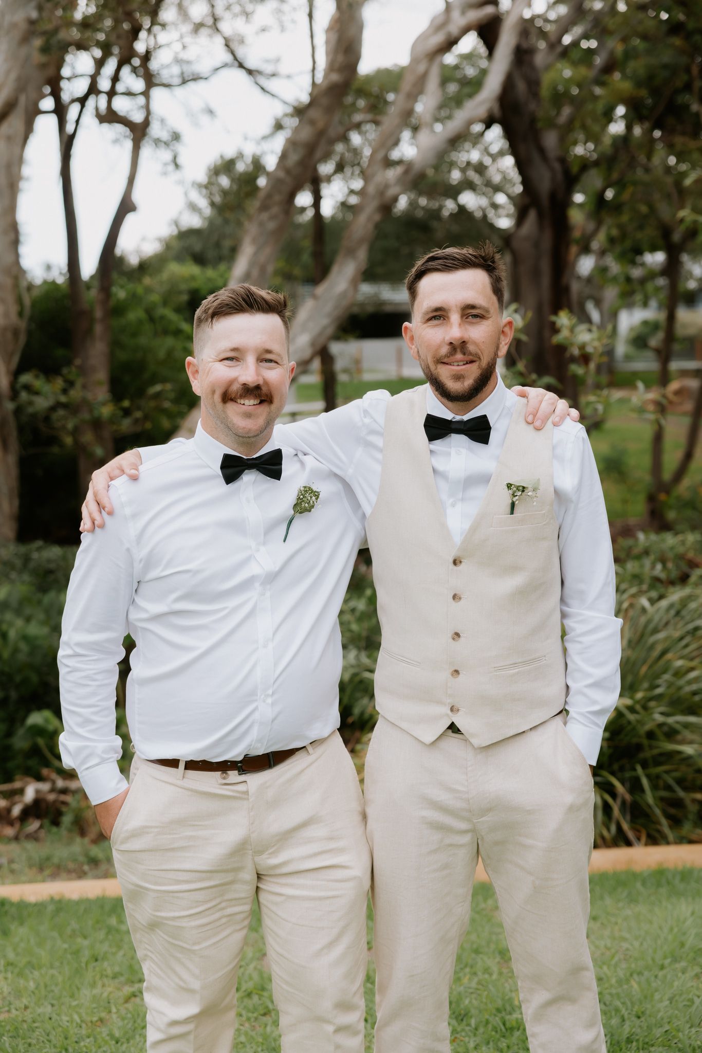 Two groomsmen in white shirts, bow ties, and beige trousers smiling together outdoors at a wedding.