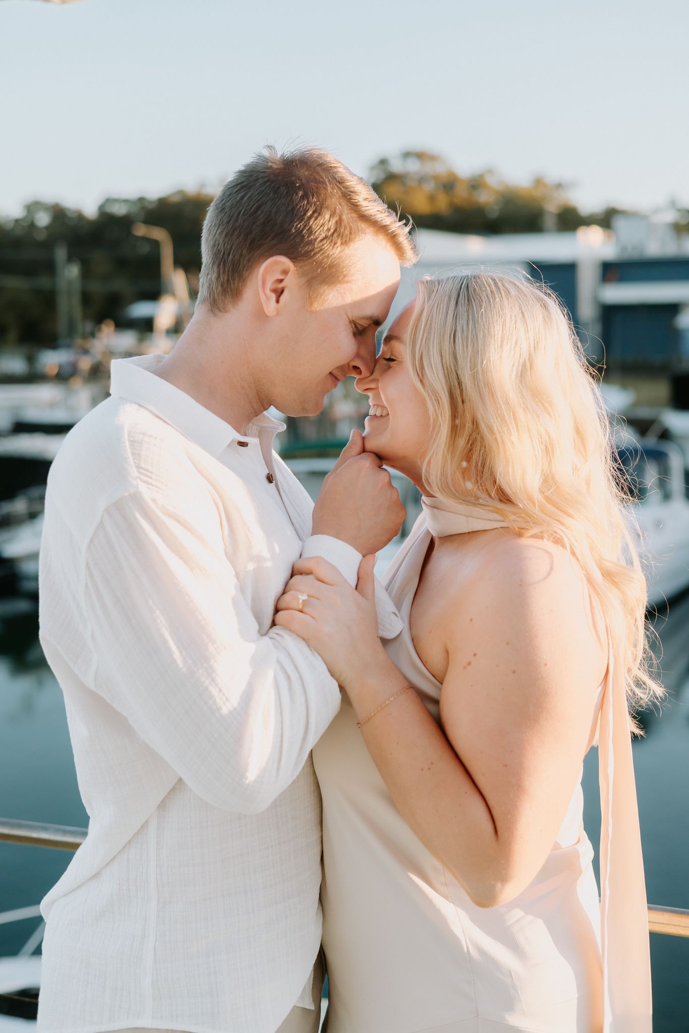 Couple in white outfits share a romantic moment on a marina dock at golden hour.