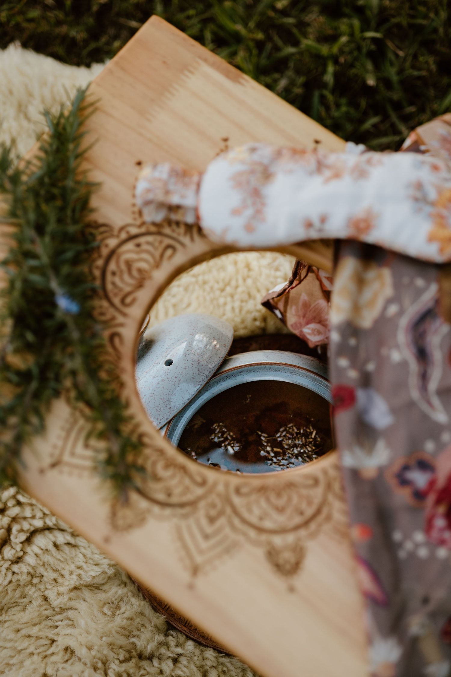 Wooden board with mandala engraving holding a ceramic teapot on a cozy blanket, surrounded by pine branches outdoors.