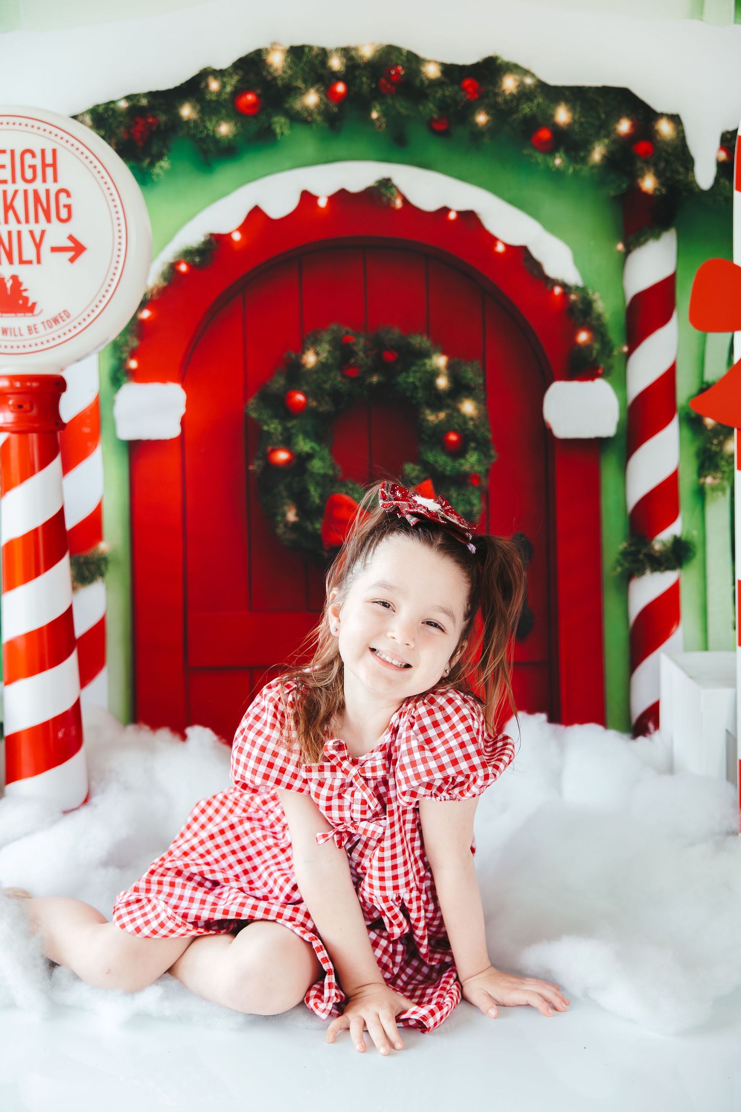Young girl in red gingham dress smiles in front of festive Christmas backdrop with candy canes and wreath.