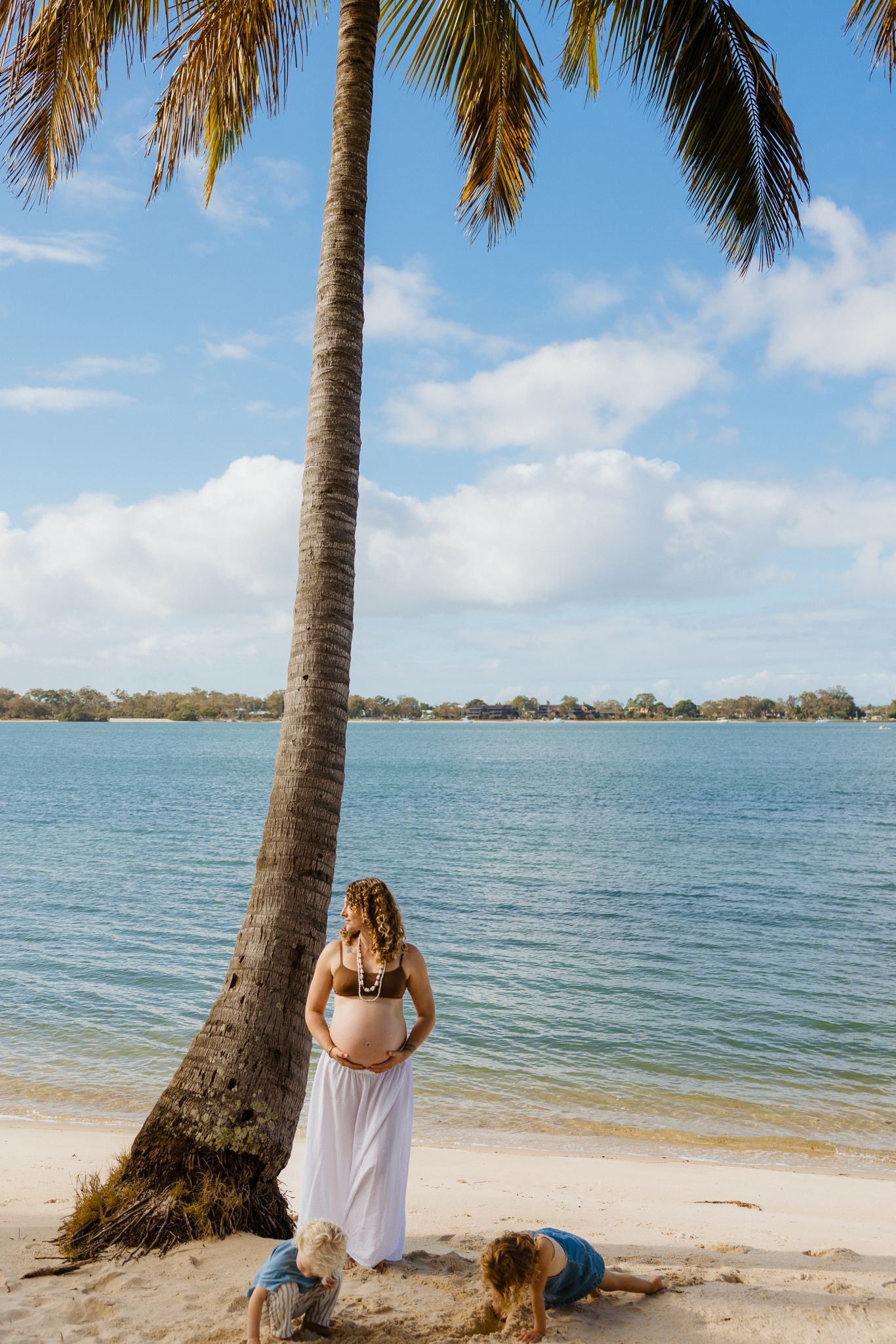 Pregnant woman in white skirt stands by palm tree on tropical beach, gazing at calm blue water with child nearby.