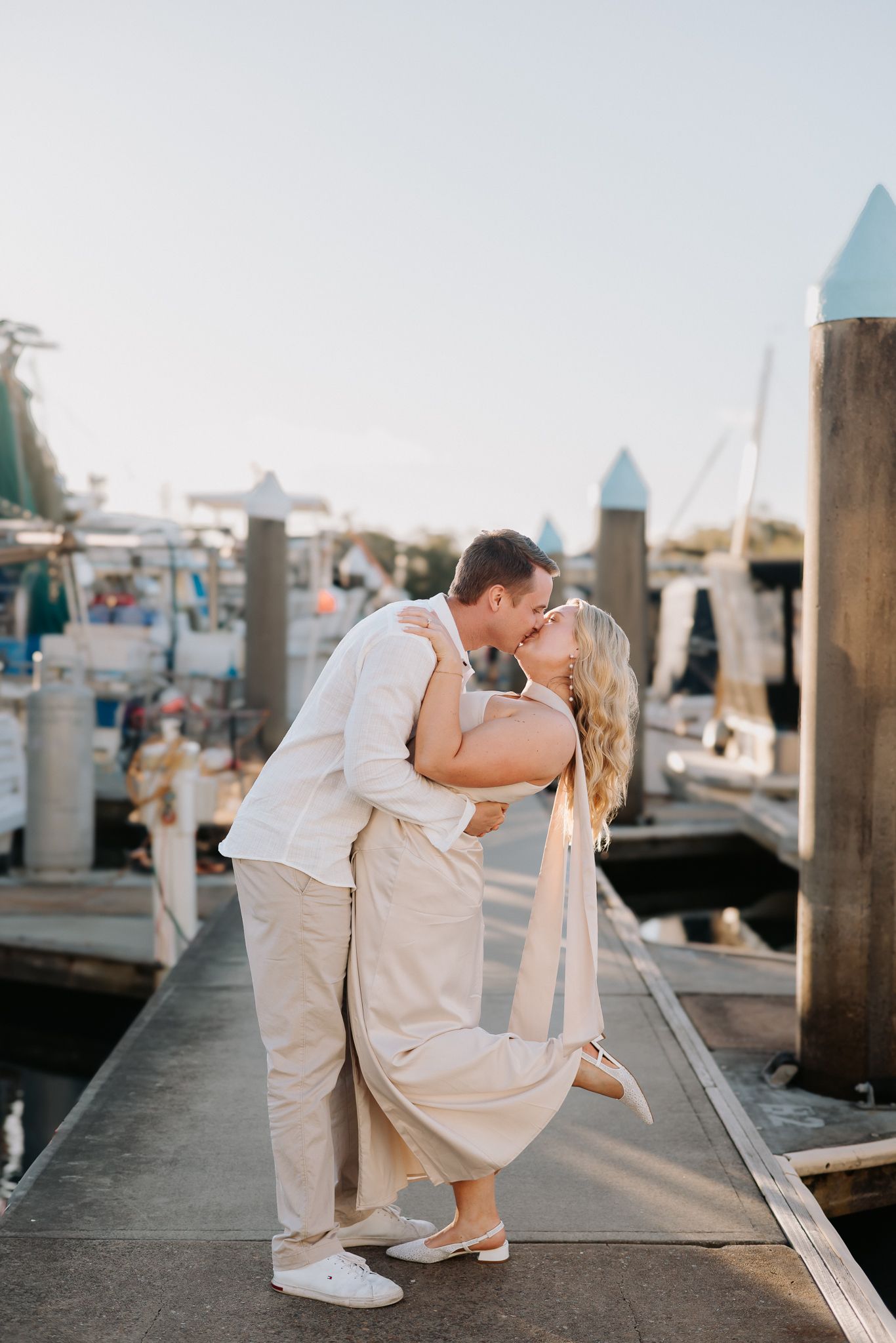 Couple sharing a romantic dip kiss on a marina dock at golden hour, dressed in elegant cream outfits.