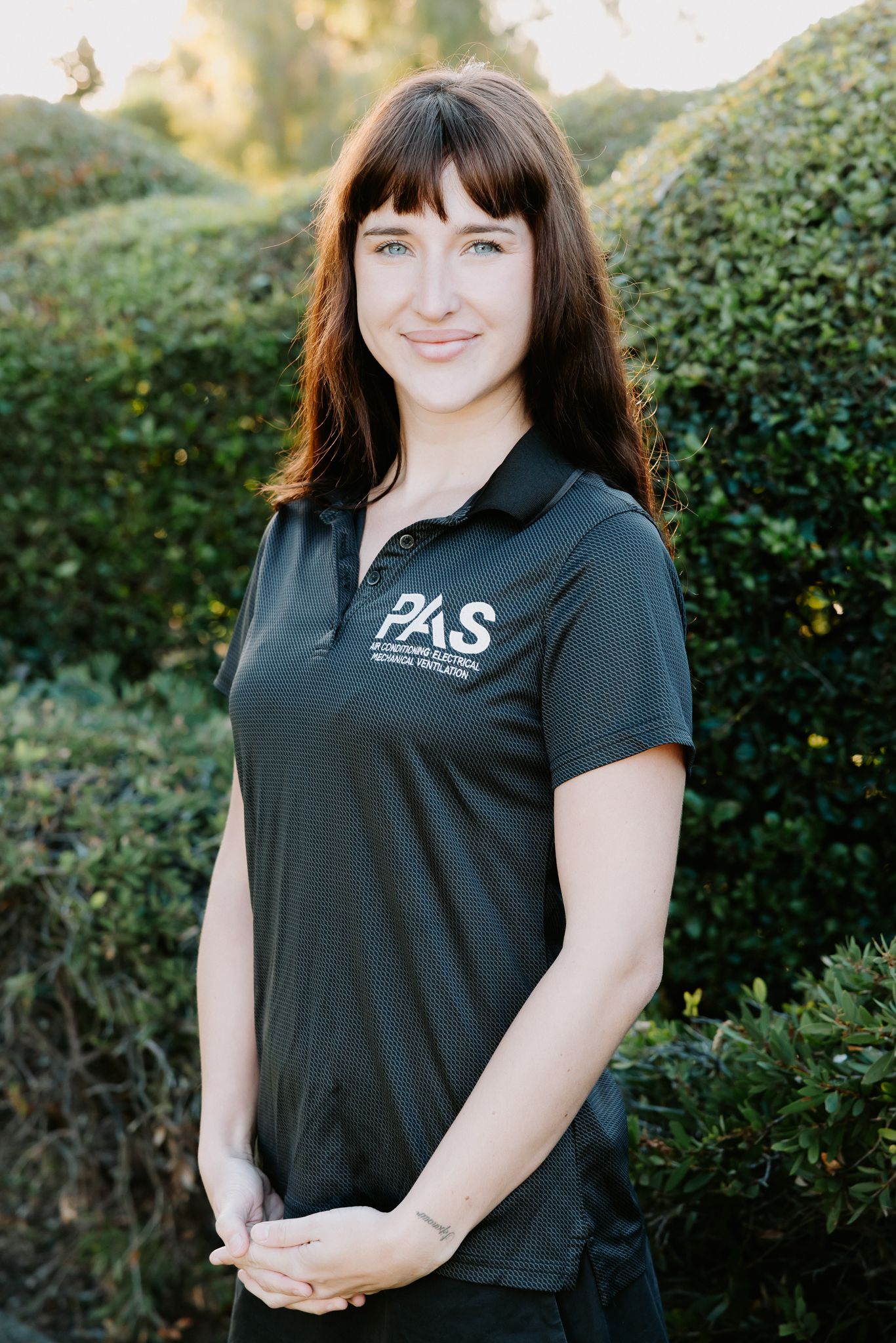 PAS employee in branded black polo shirt smiling outdoors with green hedges in background.