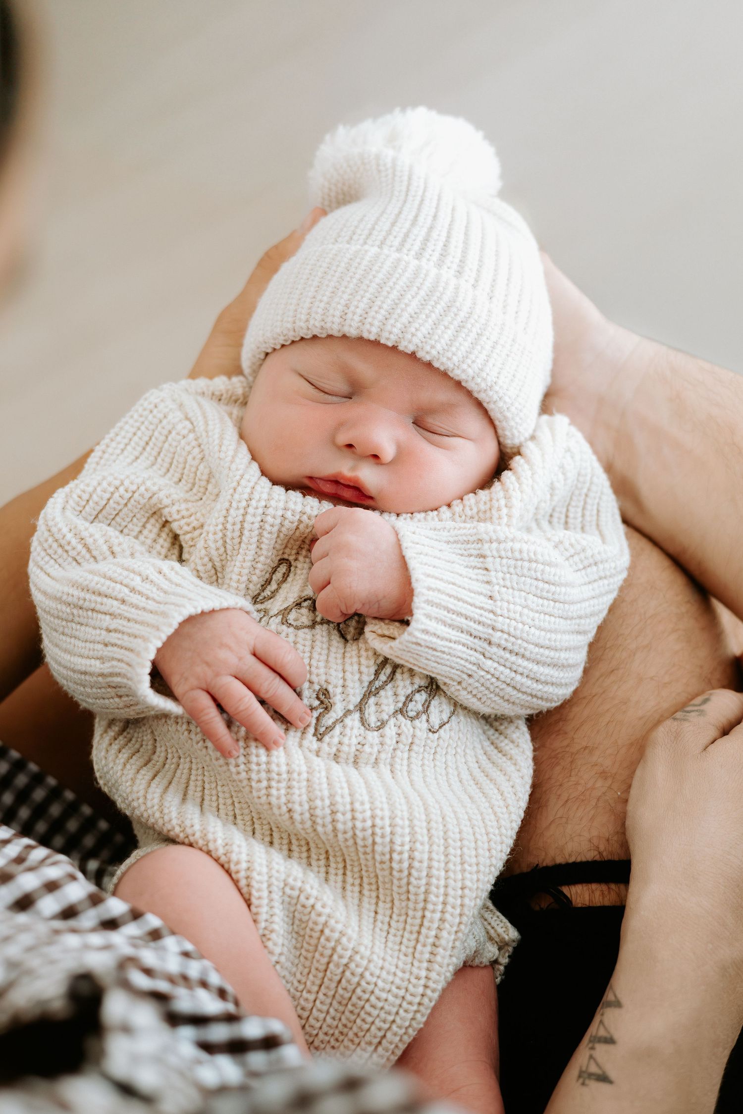 Sleeping newborn baby in cream knit sweater and pom-pom hat cradled in parent's arms.