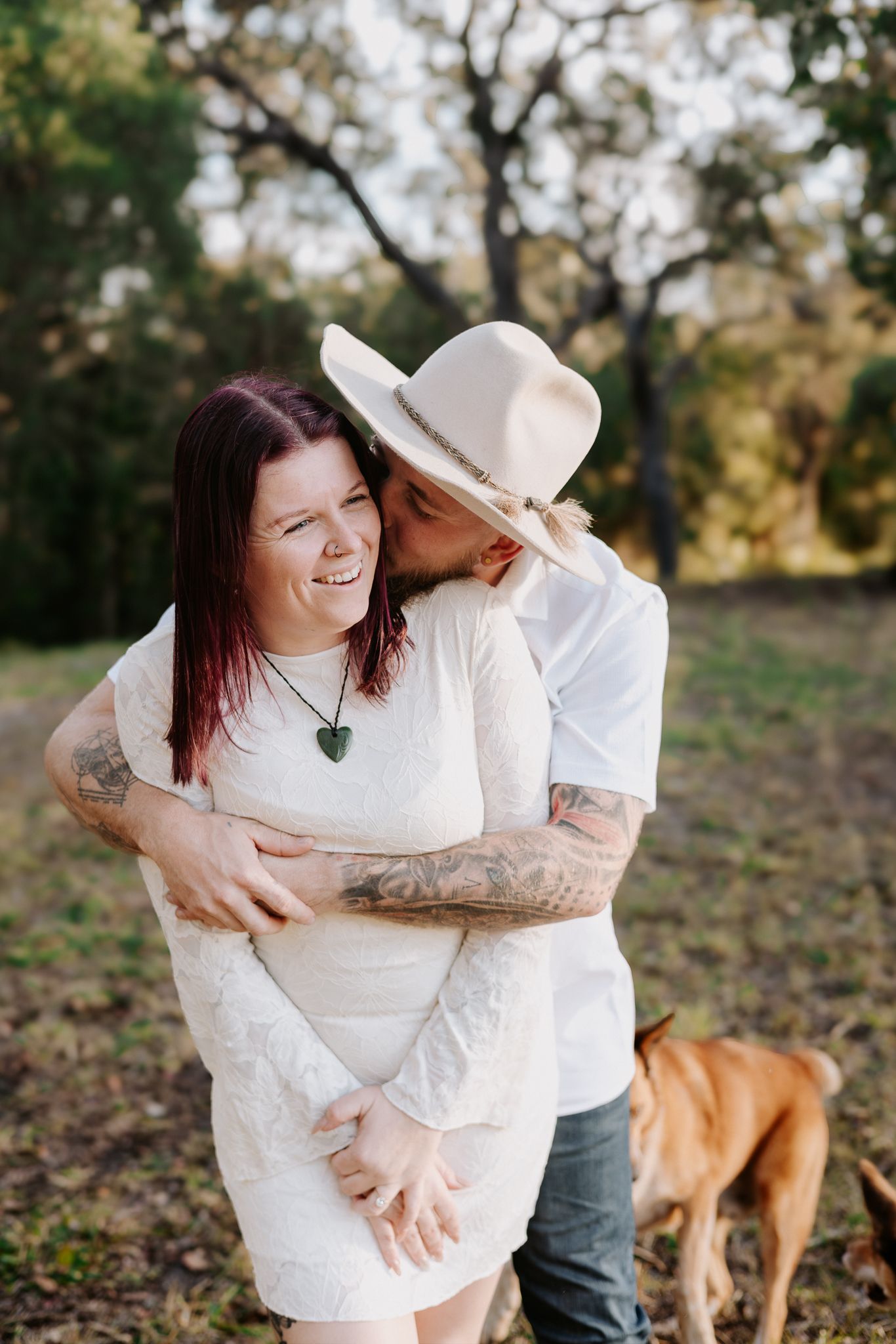 Tattooed couple embracing outdoors, woman in white lace dress with heart necklace, man in cream hat kissing her cheek, dog nearby.