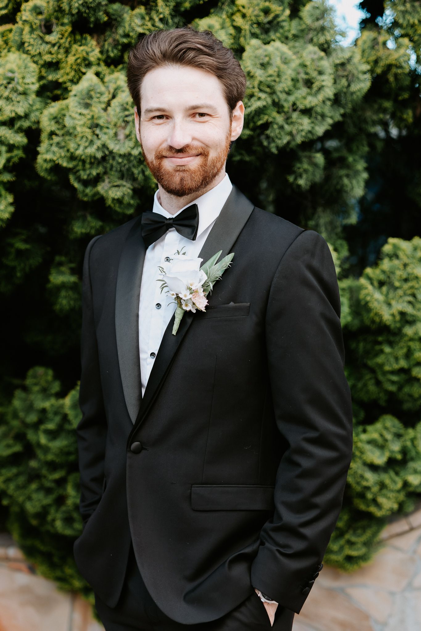 Smiling groom in black tuxedo with bow tie and floral boutonniere standing outdoors near green trees.