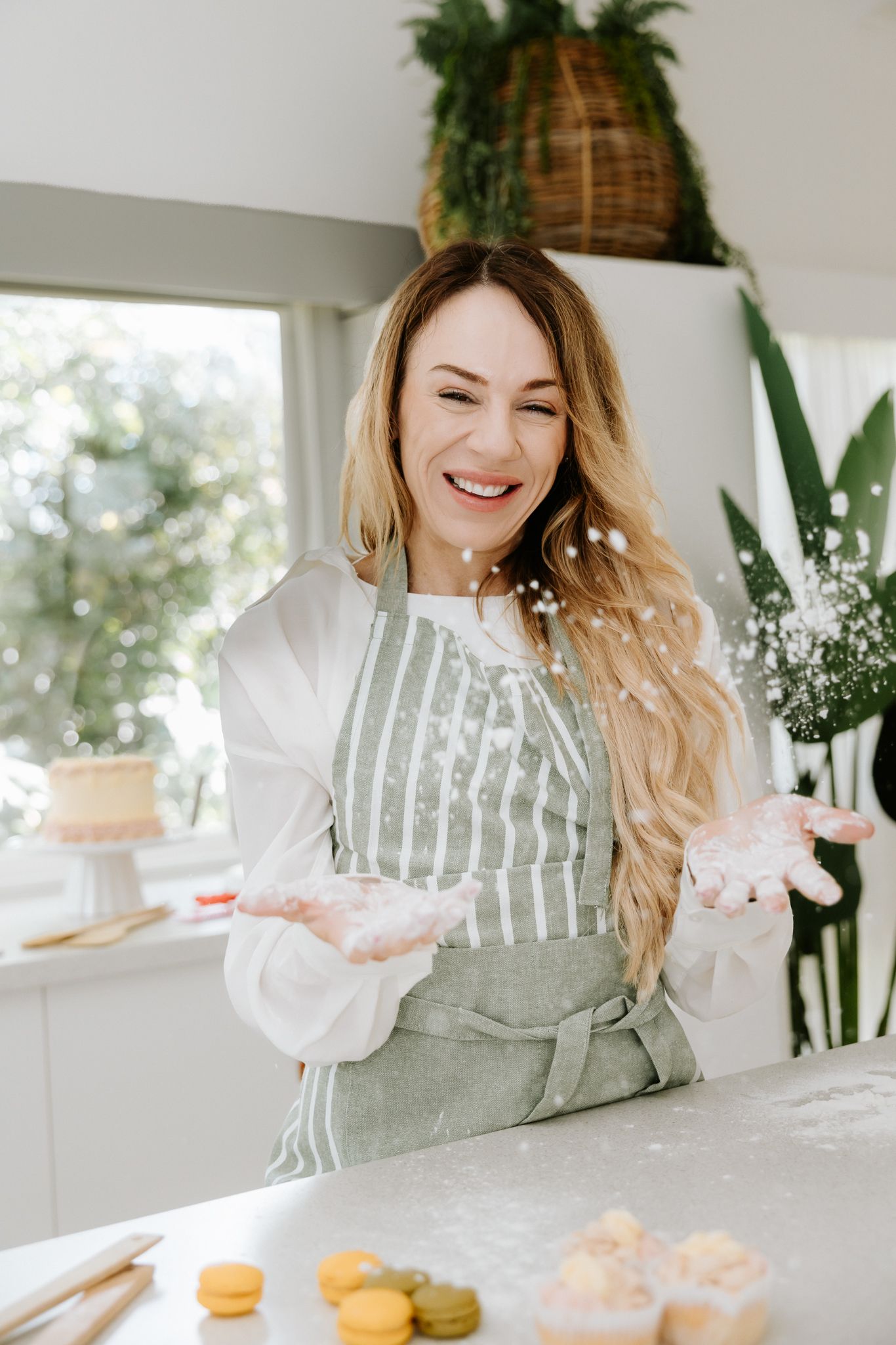 Smiling woman in striped apron clapping flour in bright kitchen with macarons and cake on counter.