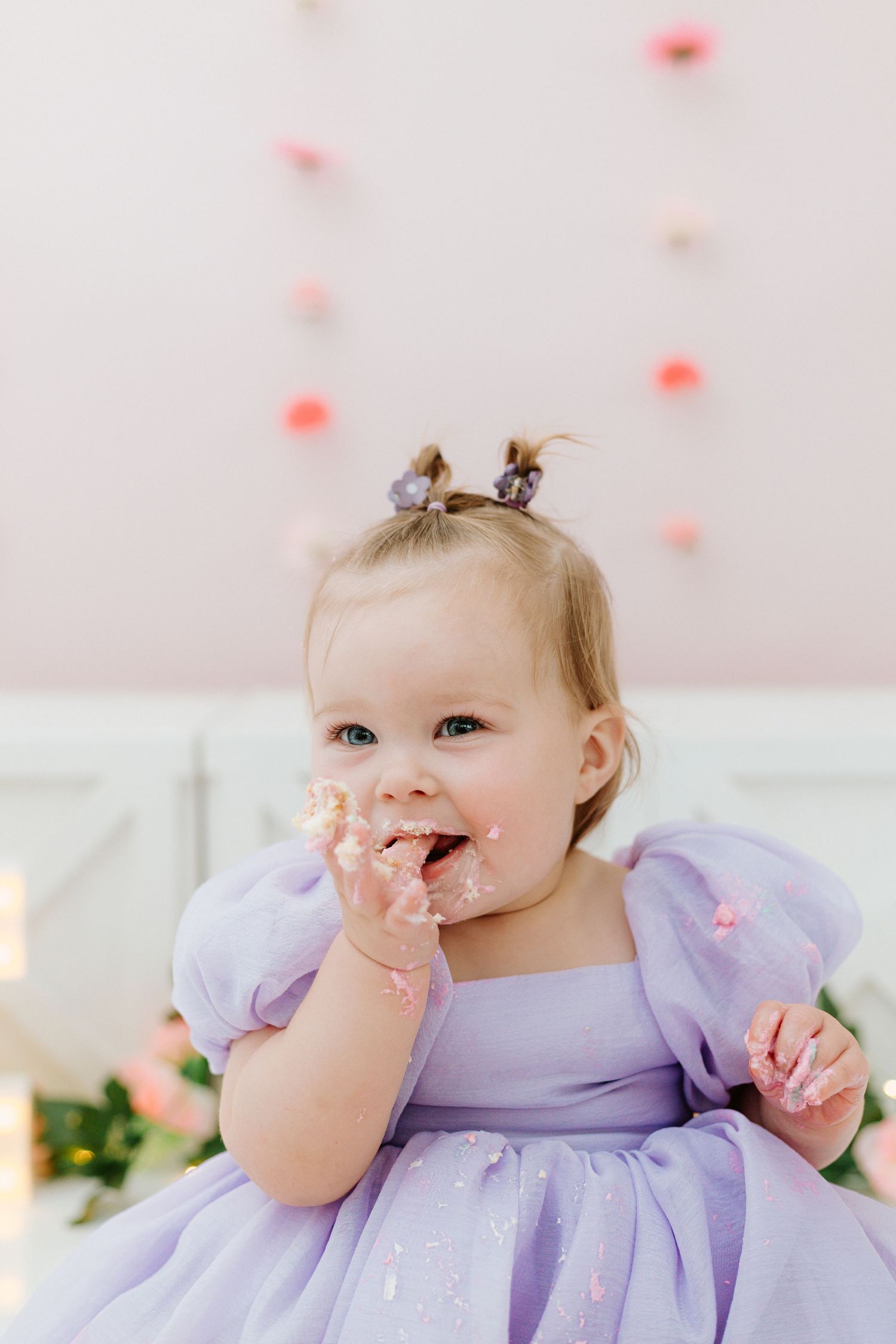 A baby in a purple dress sits surrounded by pink decorations and bokeh lights.