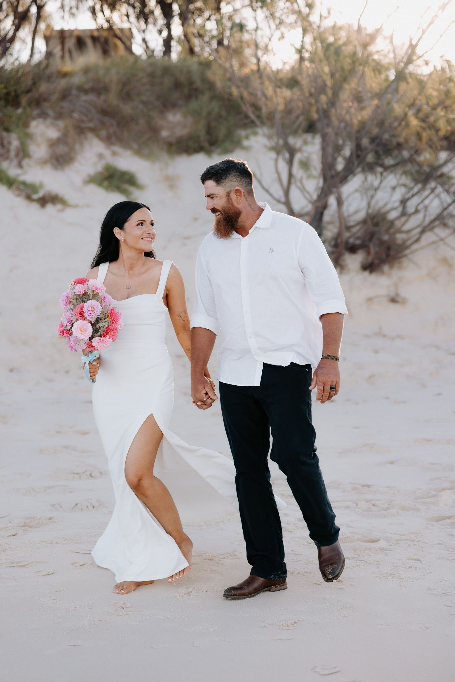 A couple walking hand in hand on sandy beach with pink bouquet.