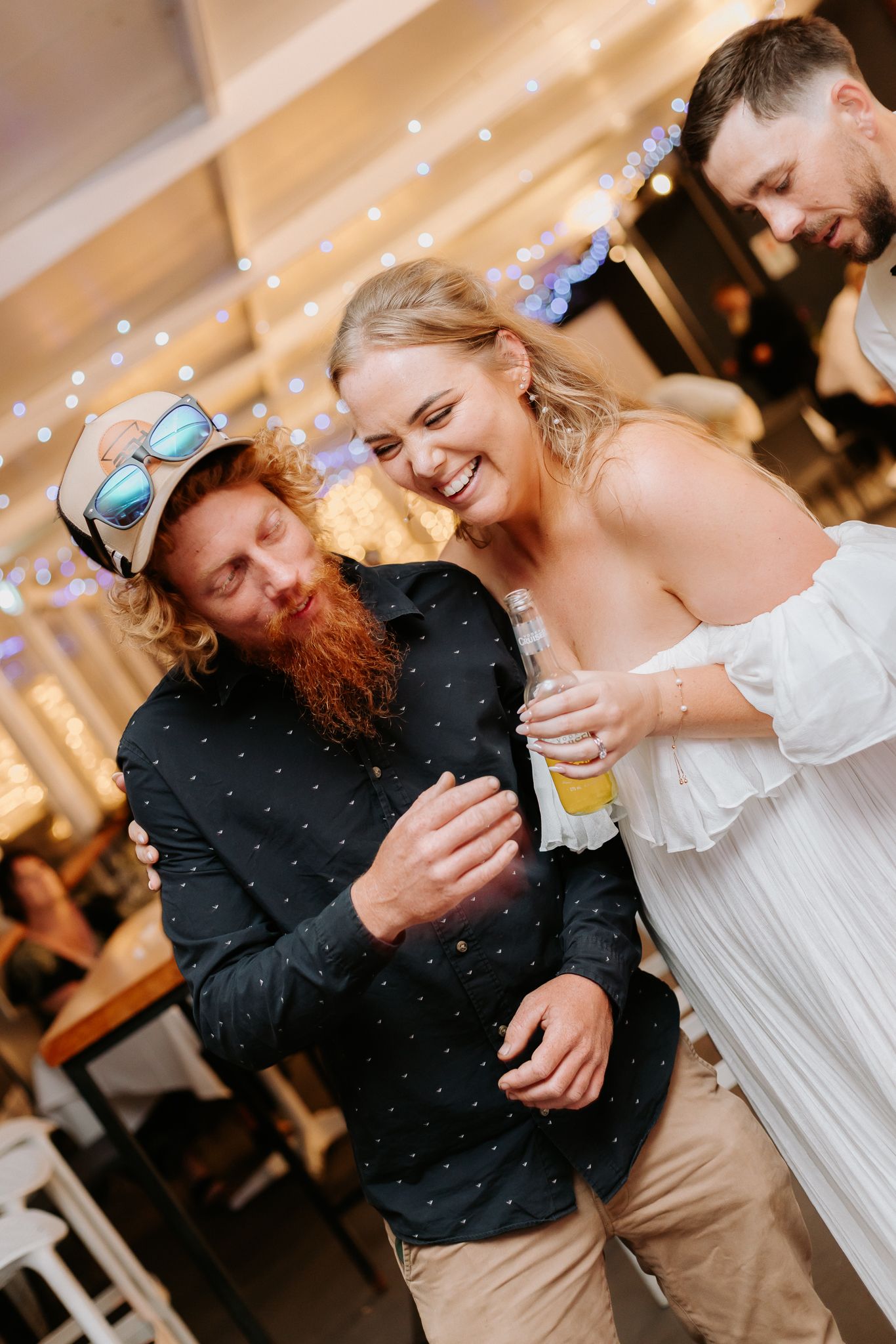Bride in white off-shoulder dress laughing with a red-bearded guest holding a beer at wedding reception.