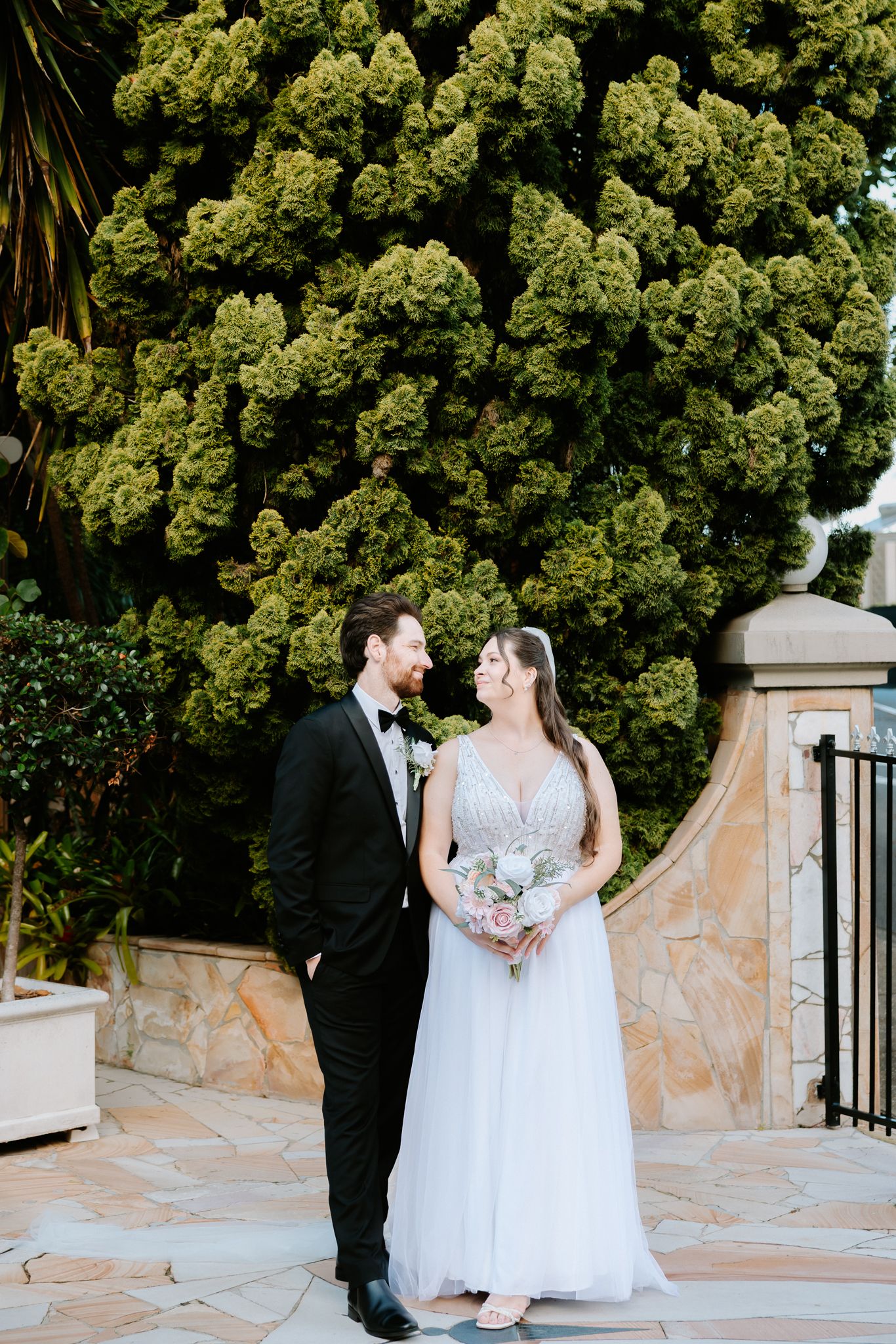 Bride and groom share a romantic moment in front of a lush green tree, bride holding a white bouquet.