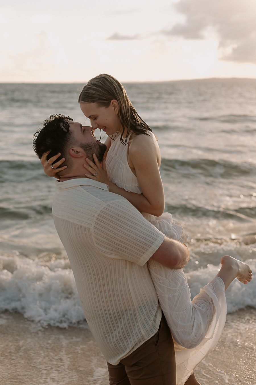 Couple sharing a romantic kiss on beach, man lifting woman in white dress amid ocean waves at sunset.
