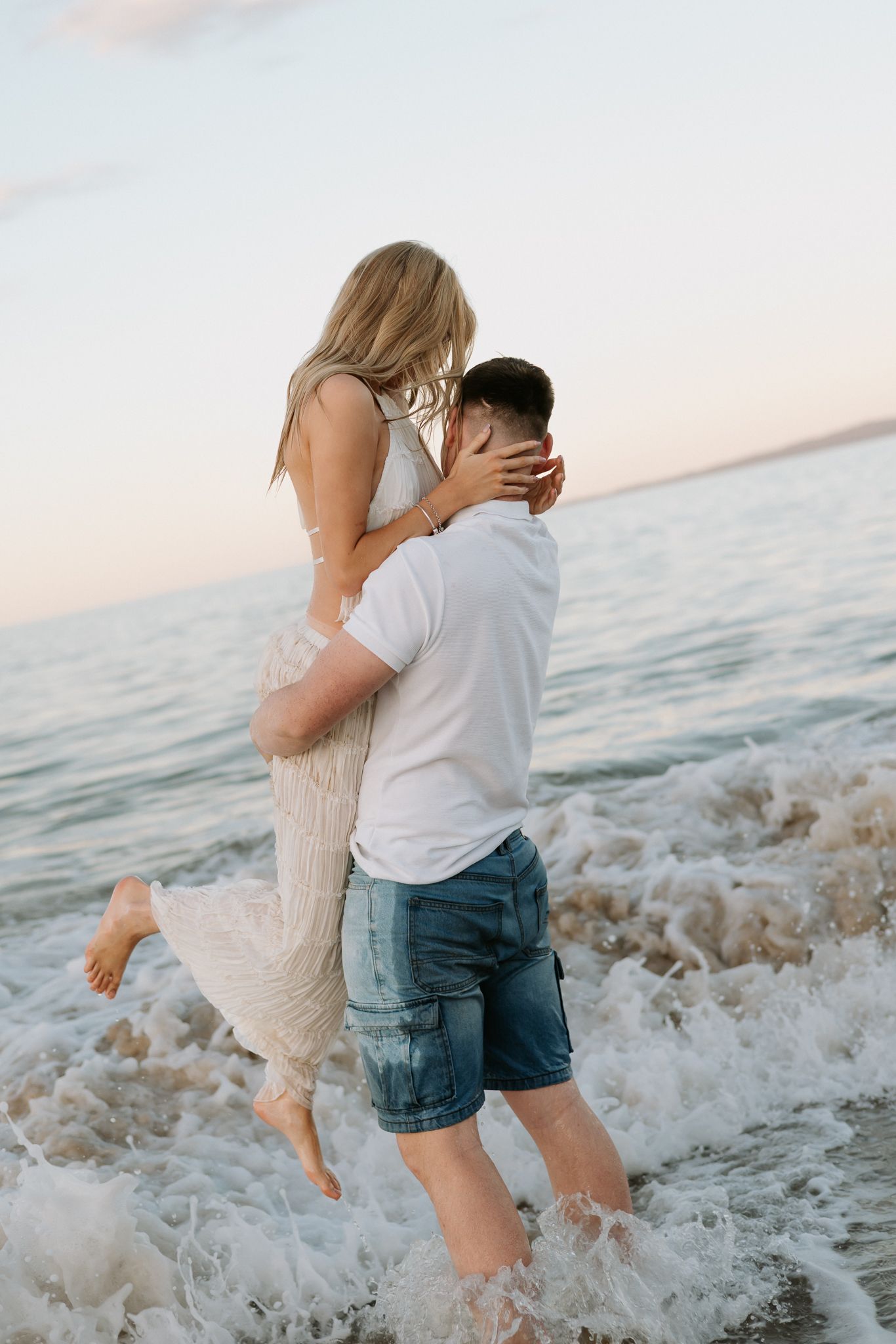 Man lifting woman in white dress while waves crash around them on beach at sunset.
