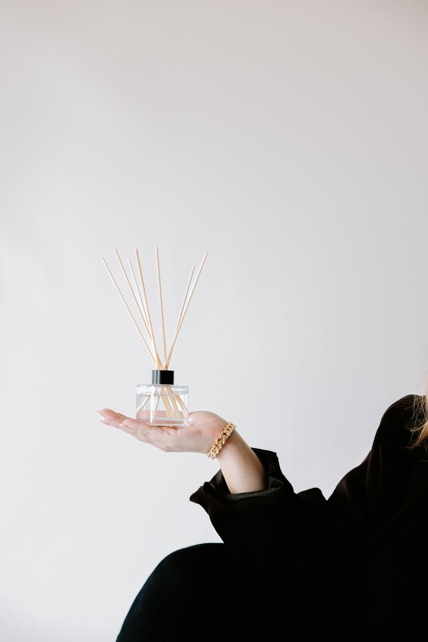 Woman in black holding a glass reed diffuser with wooden sticks on a white background.