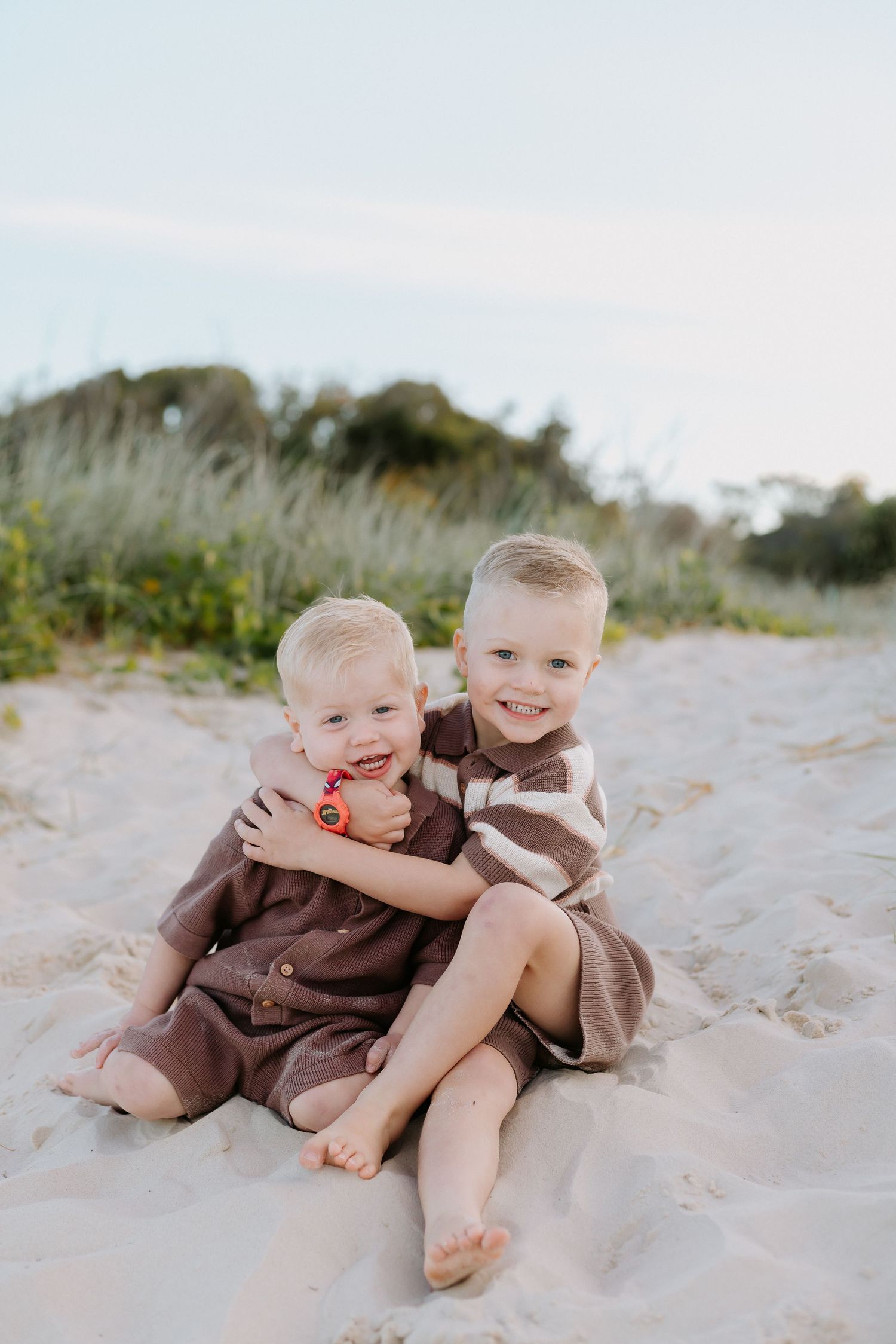 Two young blonde brothers hugging and smiling on a sandy beach, wearing brown outfits on a sunny day.