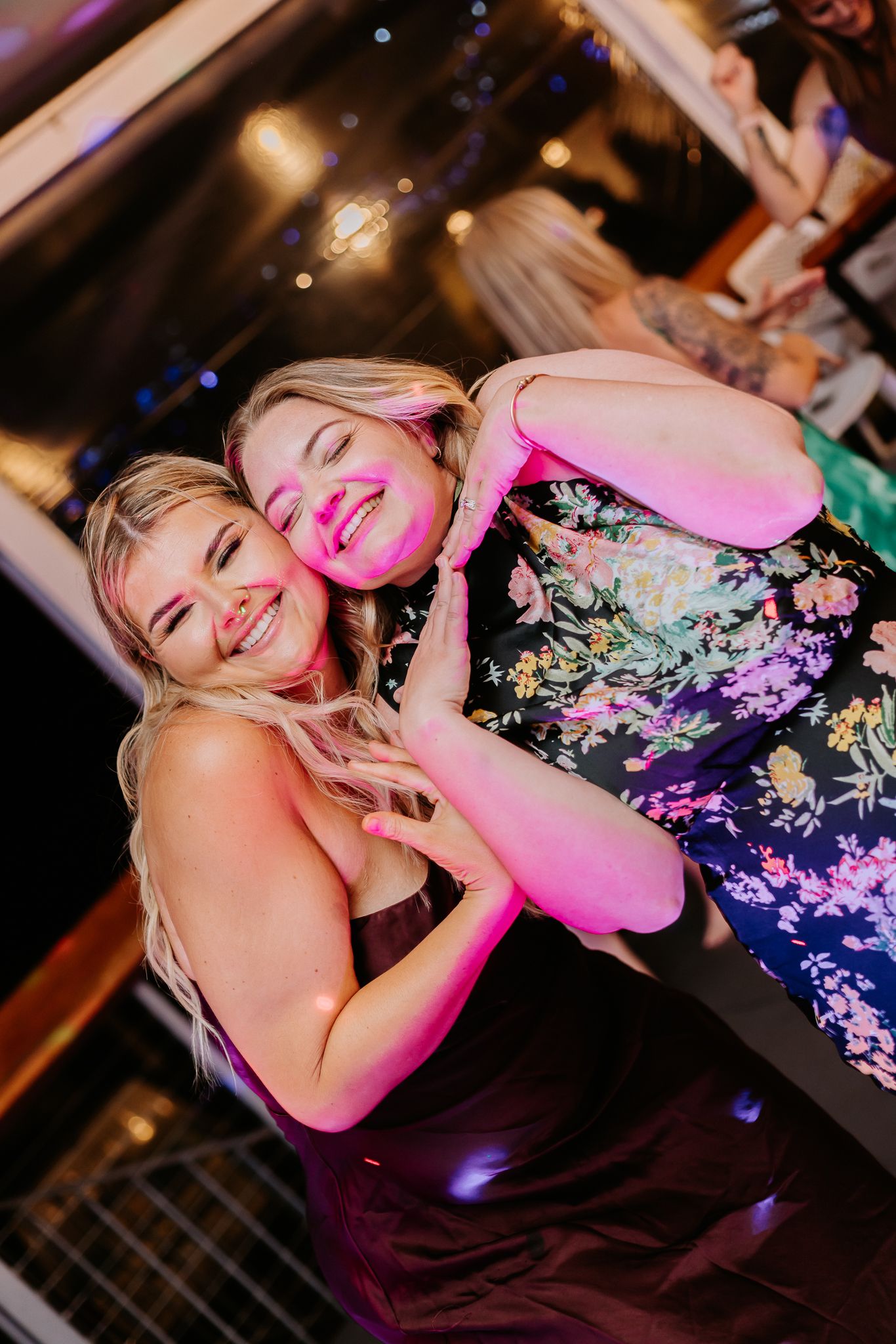 Two women in elegant dresses laughing and hugging together on a wedding reception dance floor.