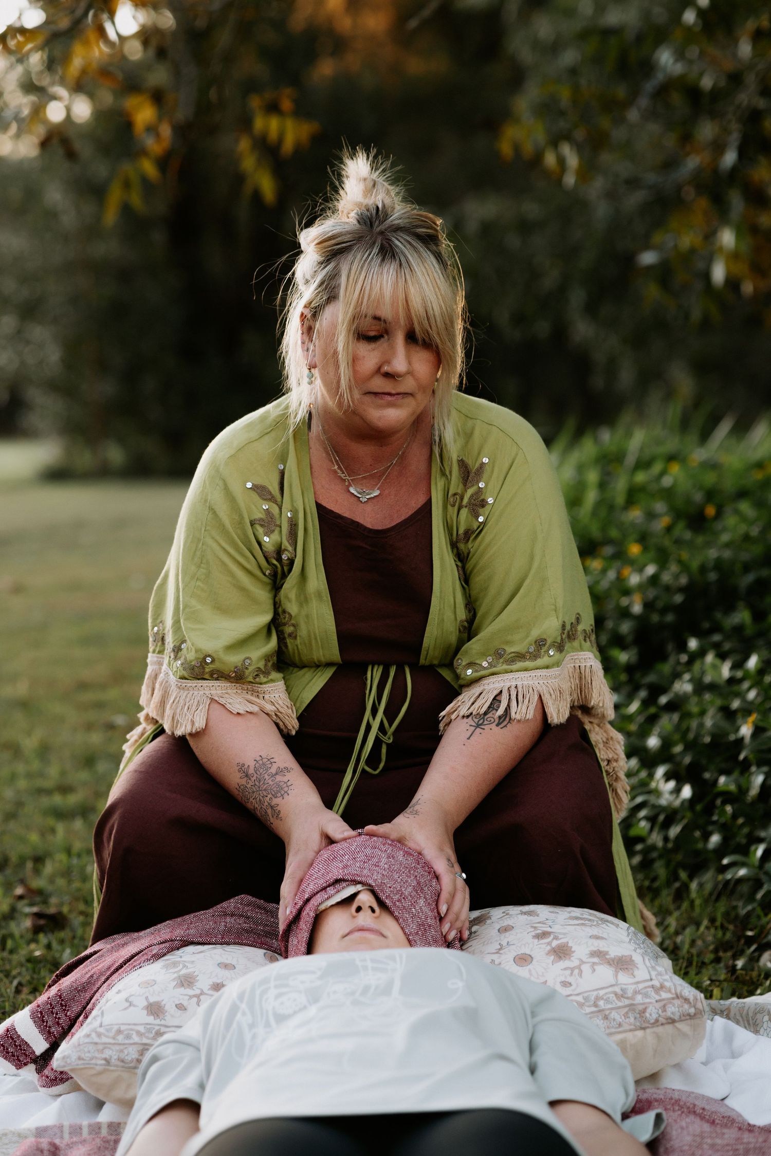 Woman in green kimono performing outdoor energy healing session on person lying on blanket in lush garden setting.