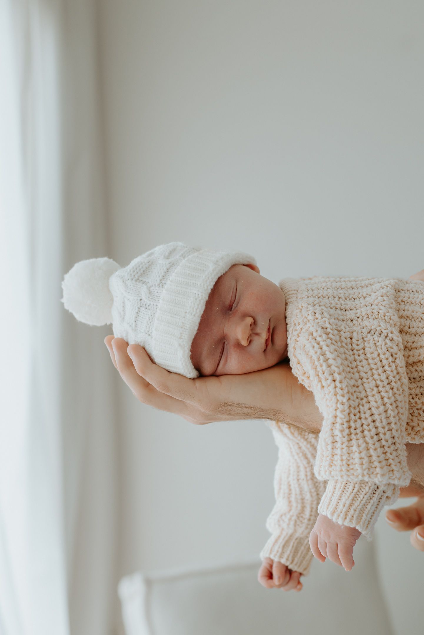 A sleeping newborn baby wrapped in a cozy white knit blanket lies peacefully in soft natural light.