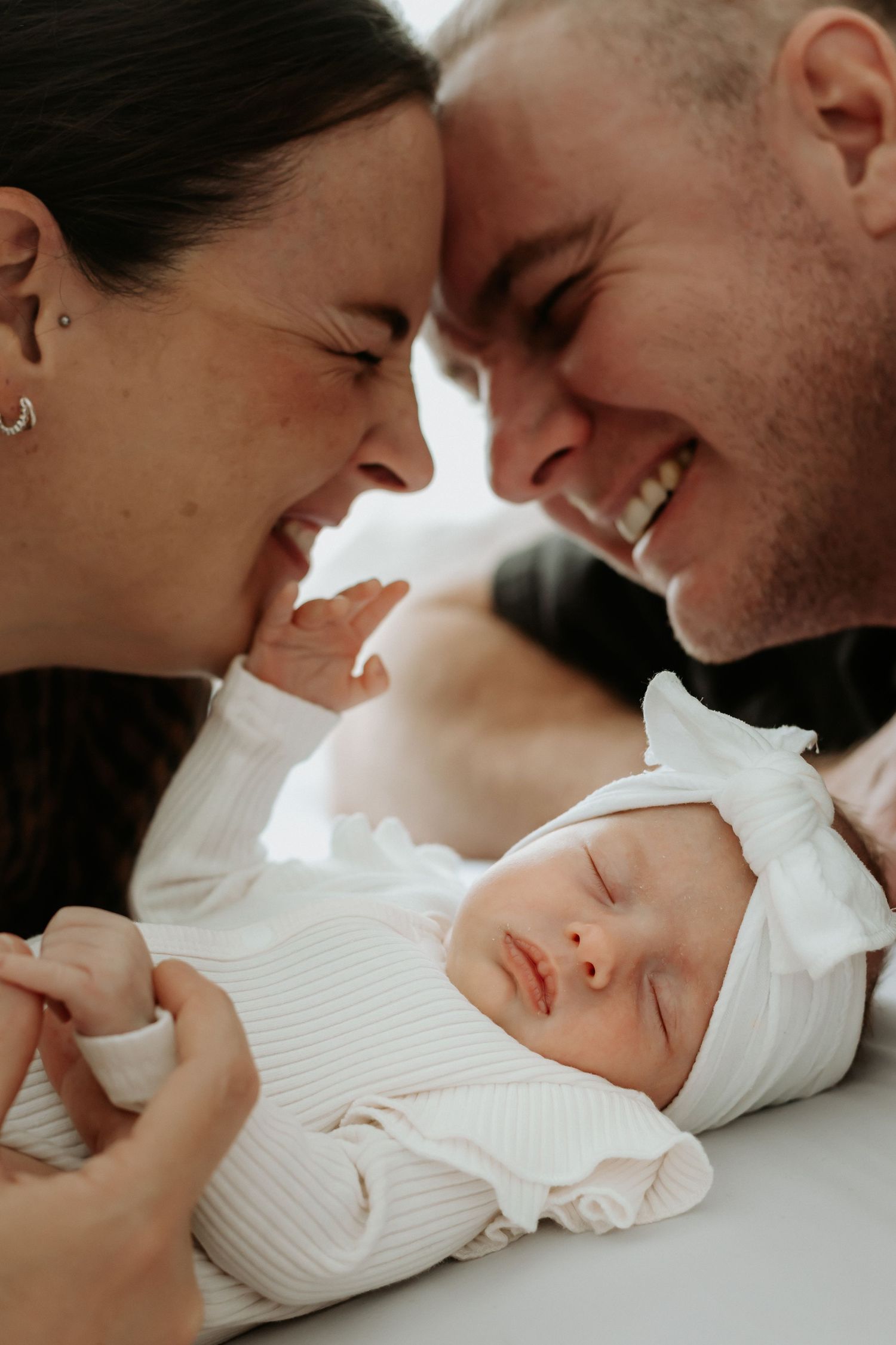 A newborn baby in white sleeps peacefully while parents share an intimate moment above.