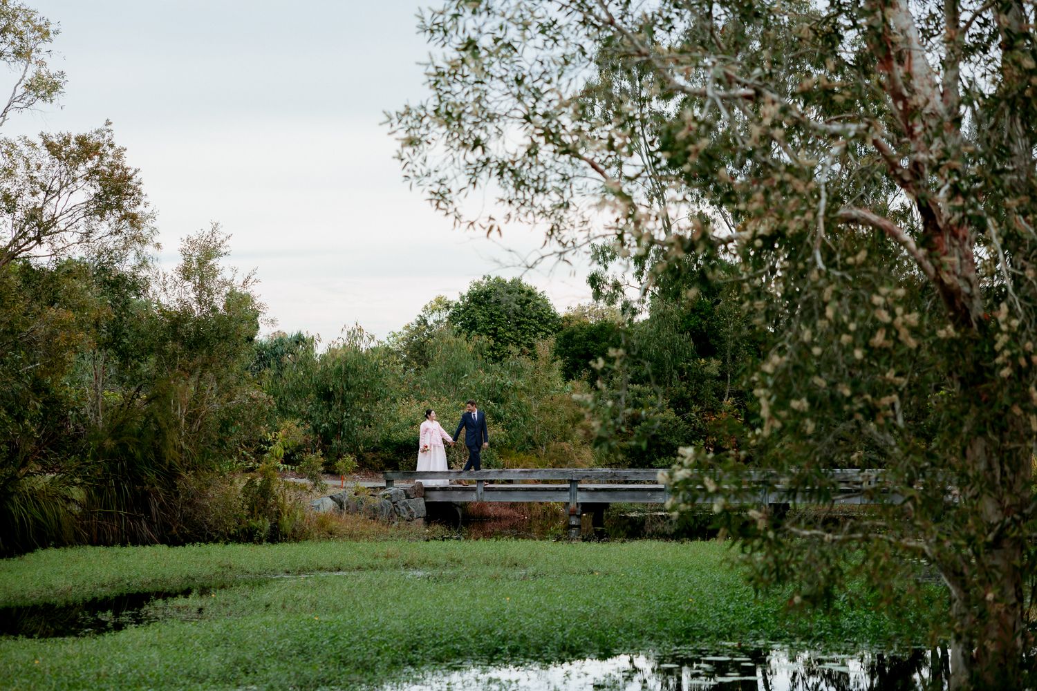 Couple standing on a wooden bridge surrounded by lush greenery and a tranquil pond in a scenic outdoor setting.