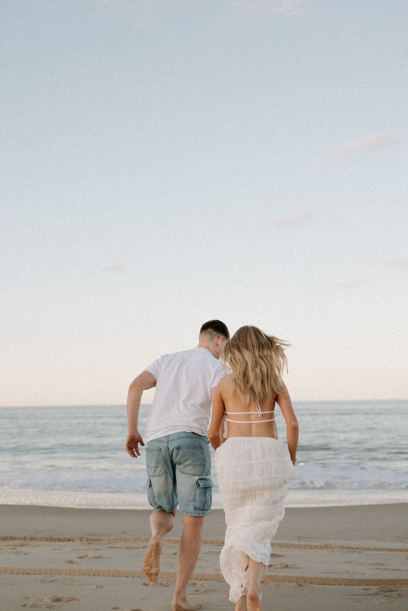 Couple running barefoot toward ocean waves on beach at golden hour, wearing white outfits.