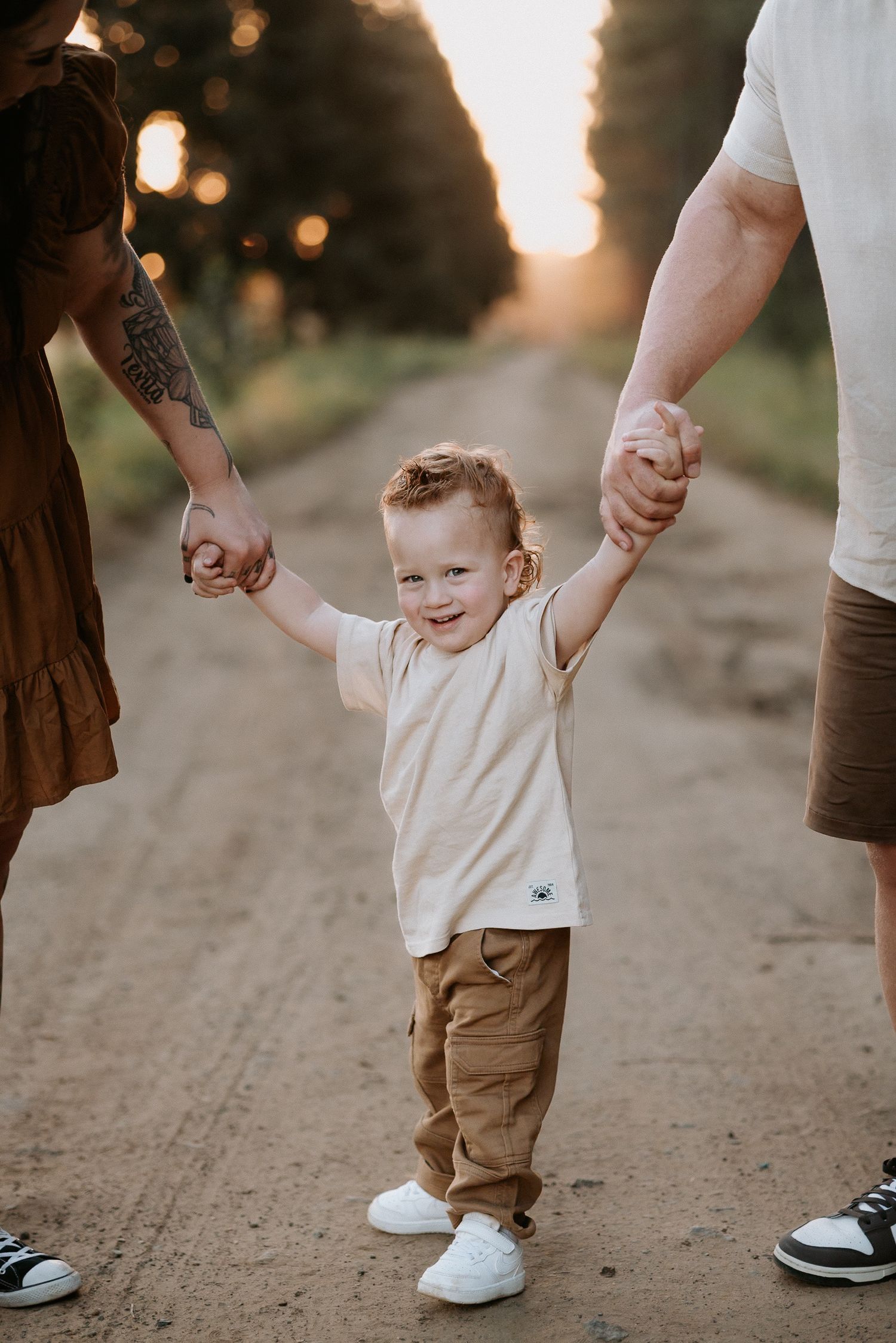 Smiling toddler boy holds hands with parents on a golden sunset dirt road, wearing a cream shirt and tan pants.