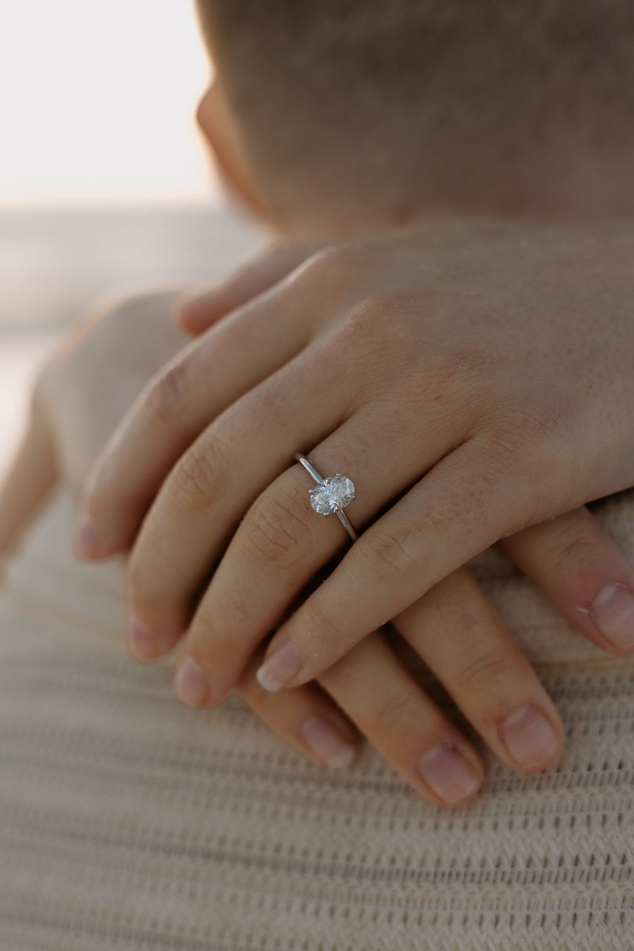 Close-up of a diamond solitaire engagement ring on a woman's finger resting on a man's shoulder.