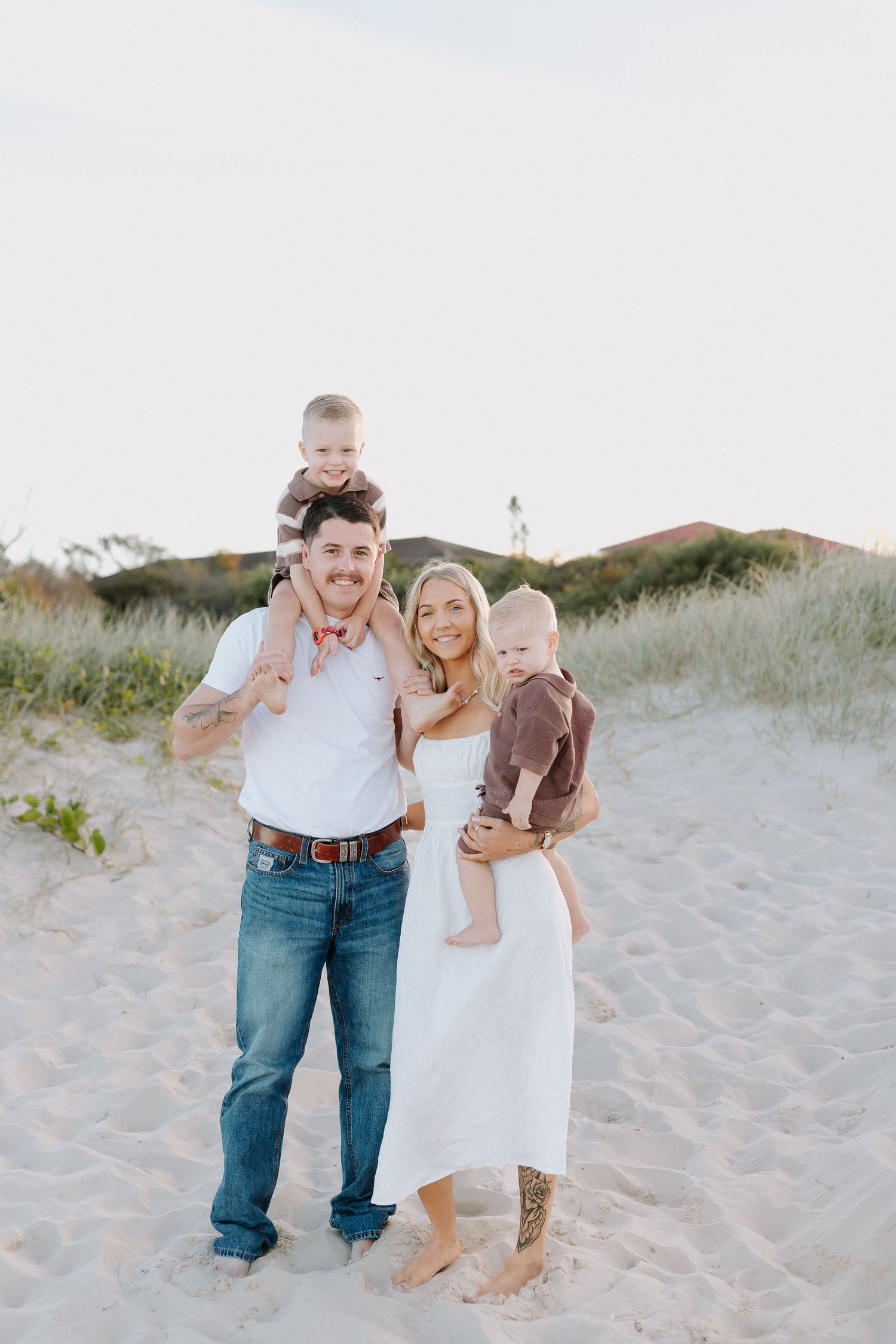 Happy family of four posing on sandy beach dunes, parents holding two young toddlers, woman in white dress.