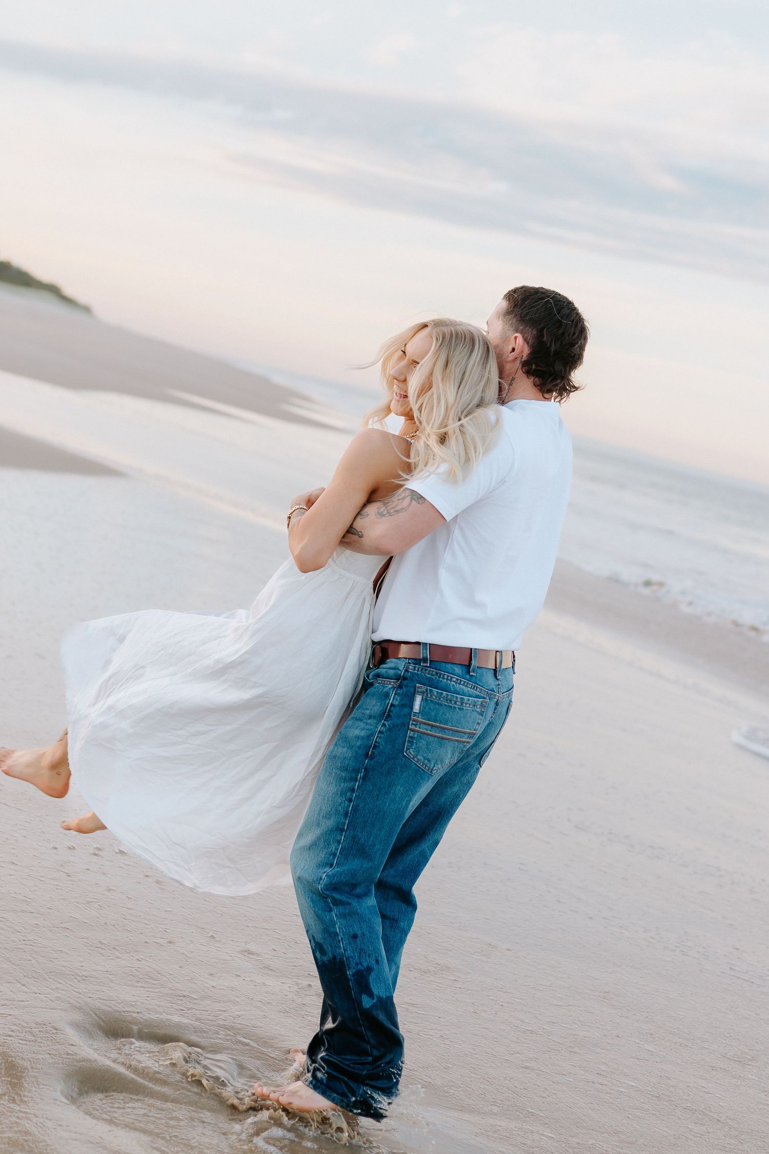 Couple embracing on a sandy beach at sunset, man lifting woman in white dress, romantic ocean backdrop.