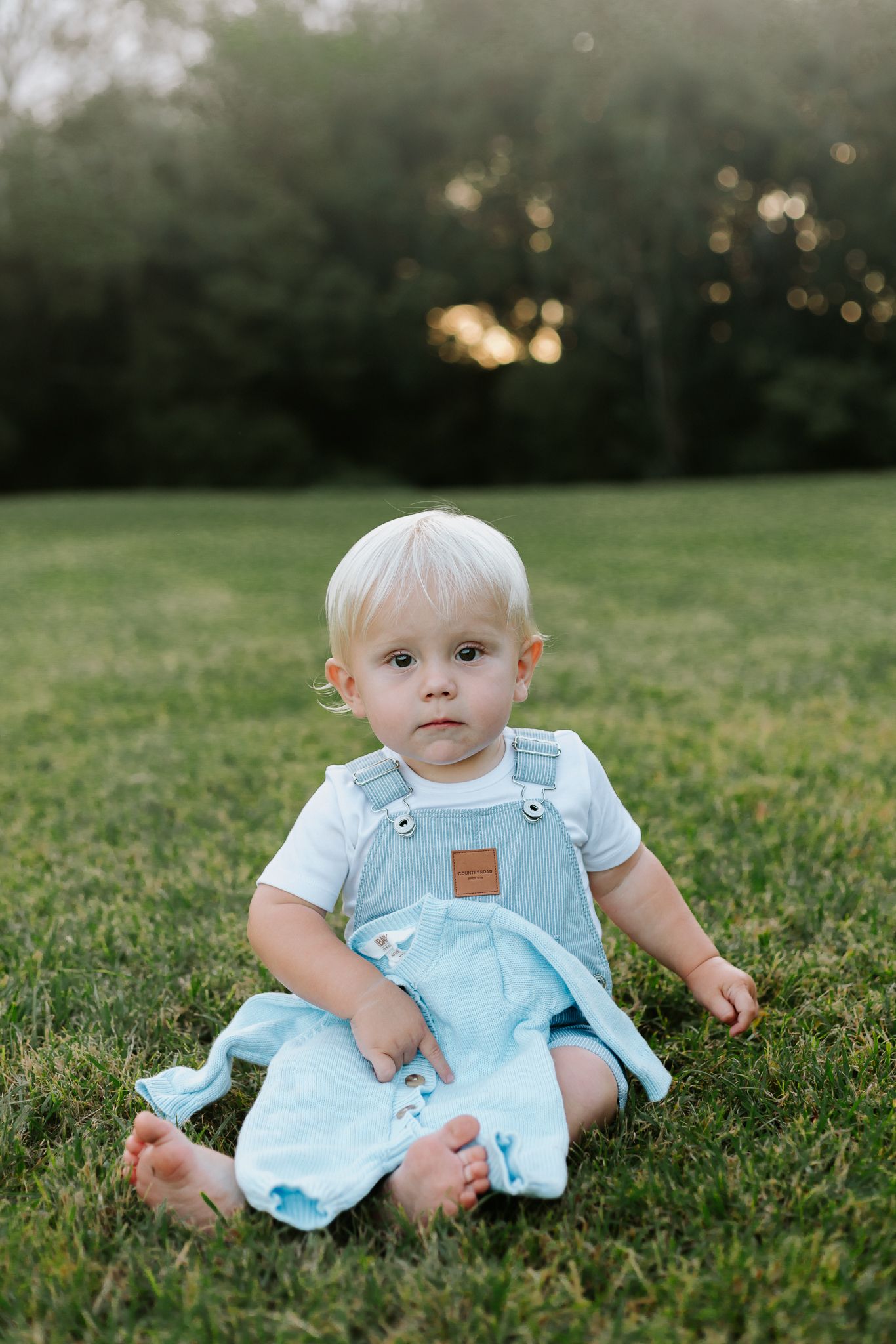 Adorable blond toddler sitting on green grass outdoors wearing light blue overalls and white shirt.
